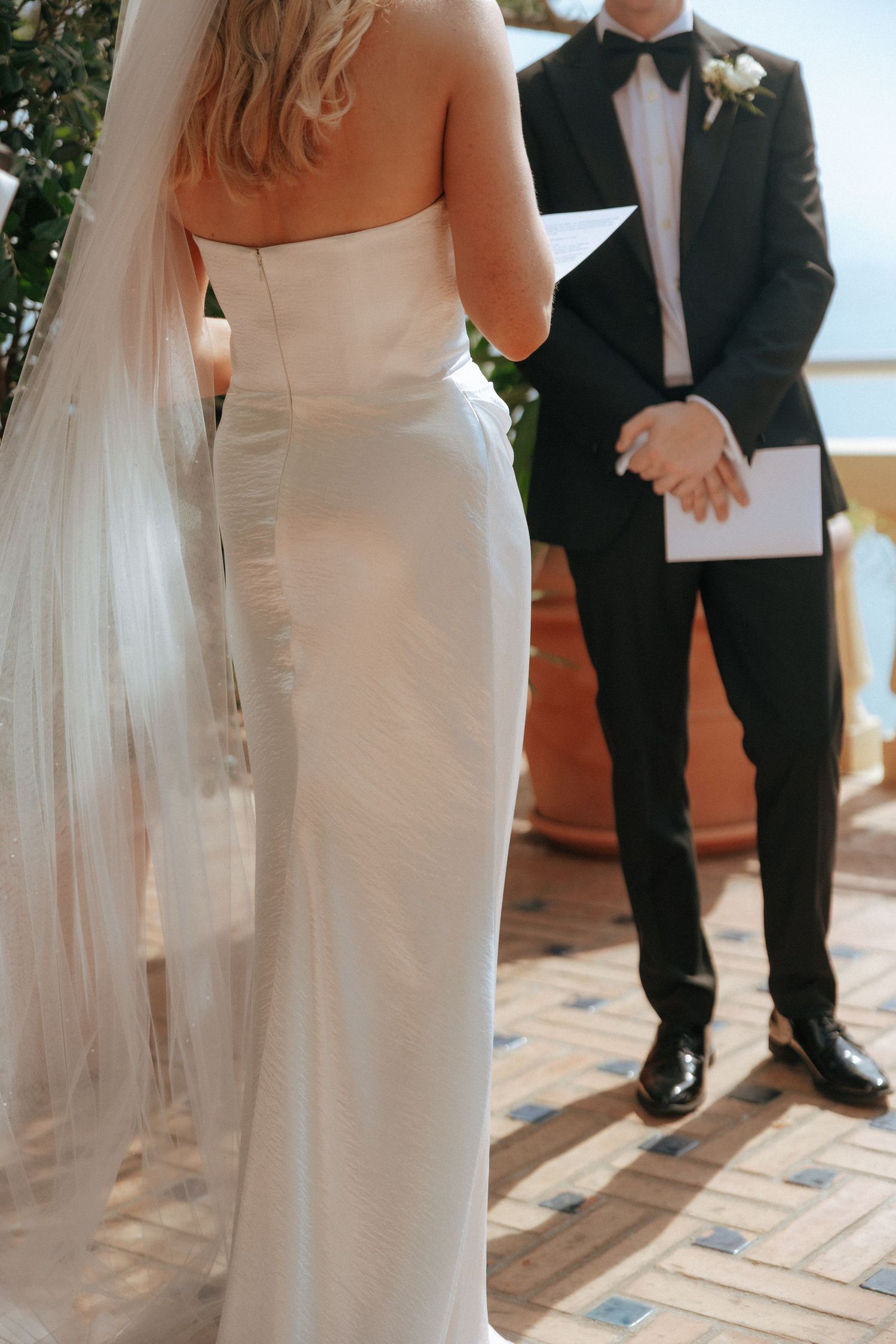 Elegant bride in fitted white dress with flowing veil on a sunlit terrace.