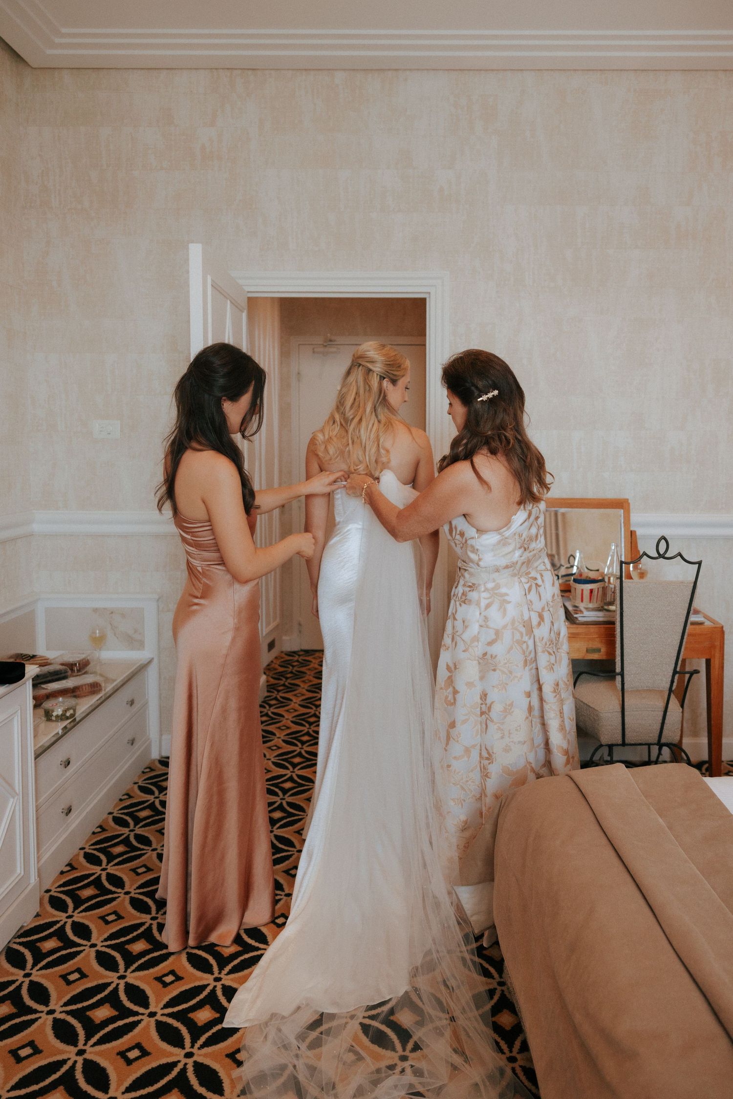 Two bridesmaids help the bride prepare on her wedding day, adjusting her flowing white gown in an elegant hotel room.