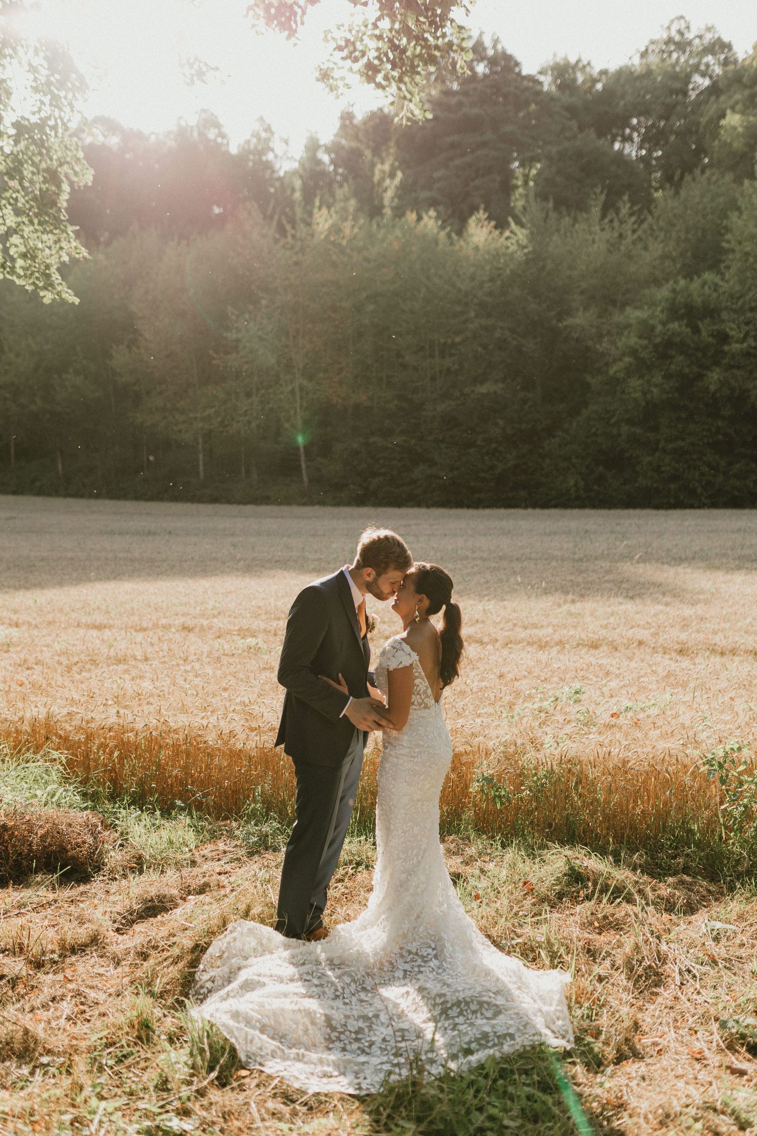 A couple in formal wear embrace in a golden sunlit field at sunset with a forest backdrop.