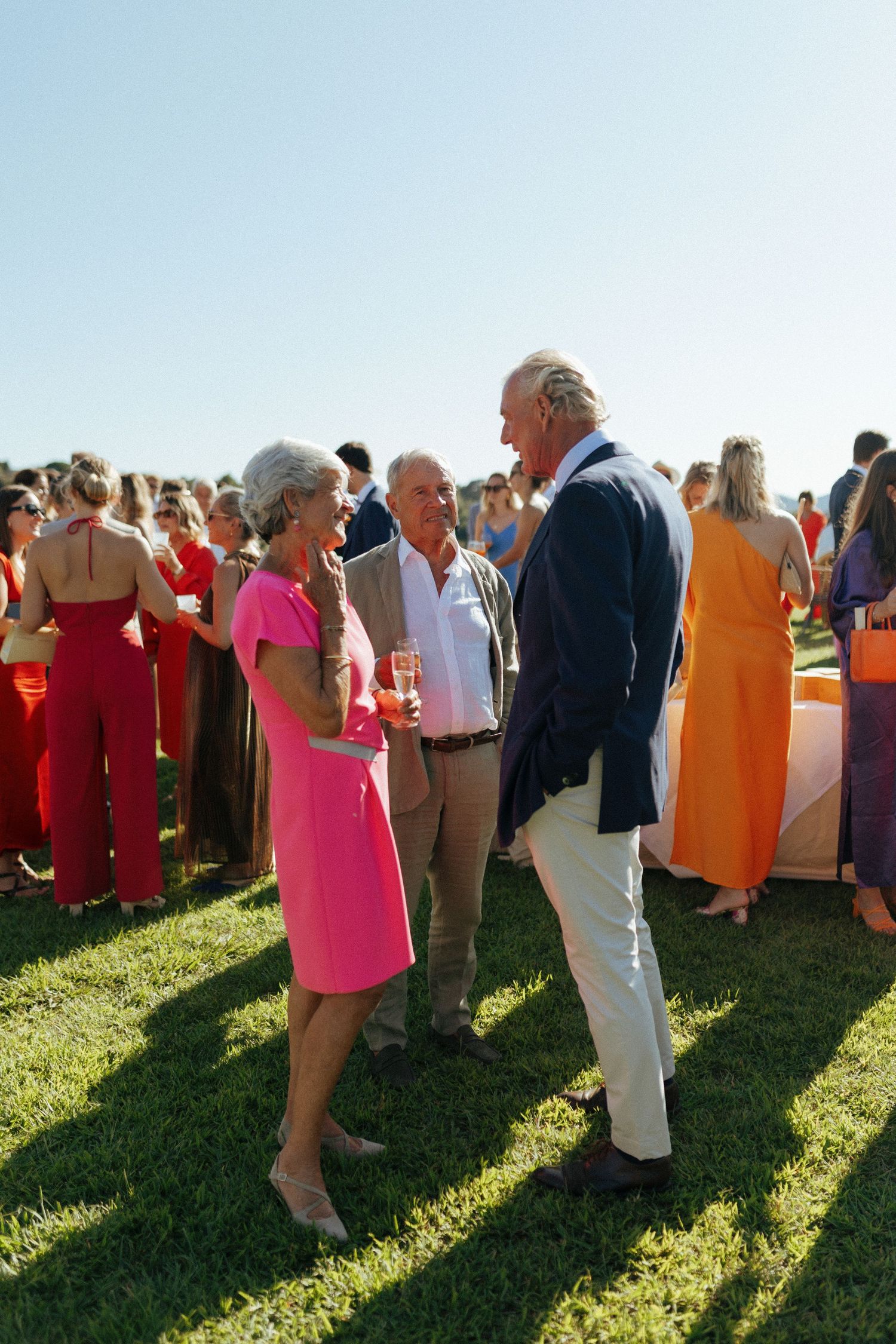 Wedding guests mingle on a grassy lawn during a sunny outdoor reception with colorful summer attire.
