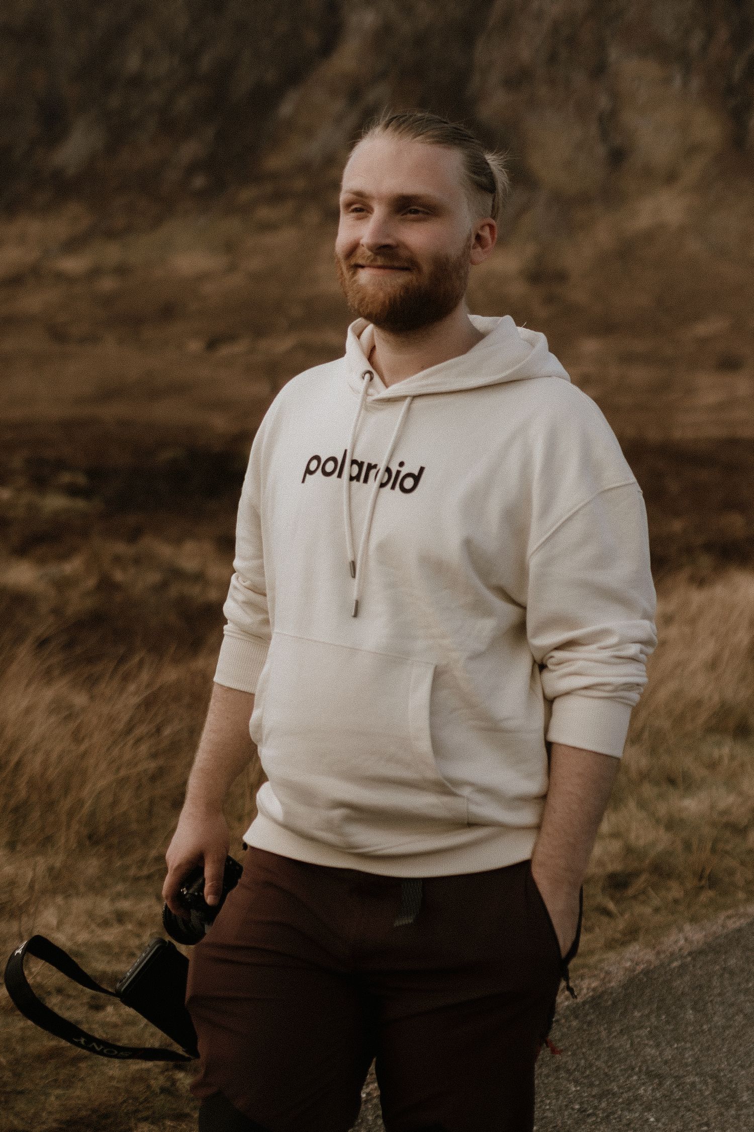 Photographer in white Polaroid hoodie and brown pants standing outdoors.