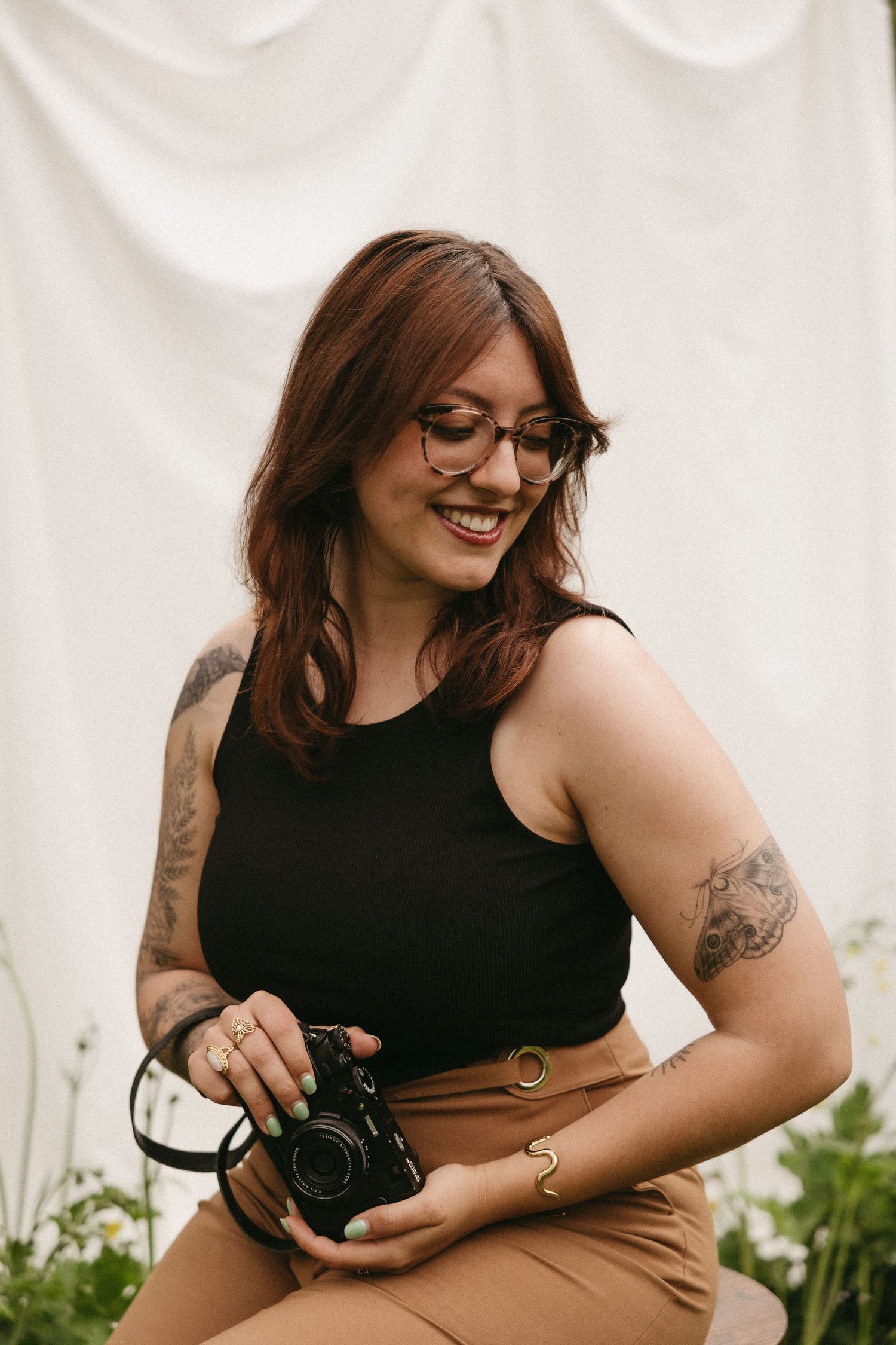 A photographer wearing glasses and a black tank smiles while holding a camera against a light backdrop.
