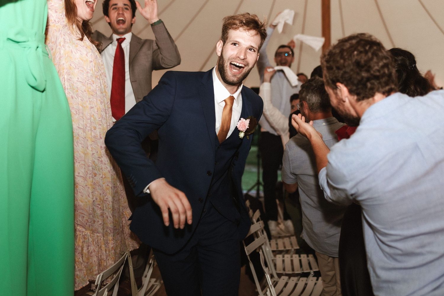 A wedding photographer captures someone in a navy suit posing on chairs during a reception celebration.