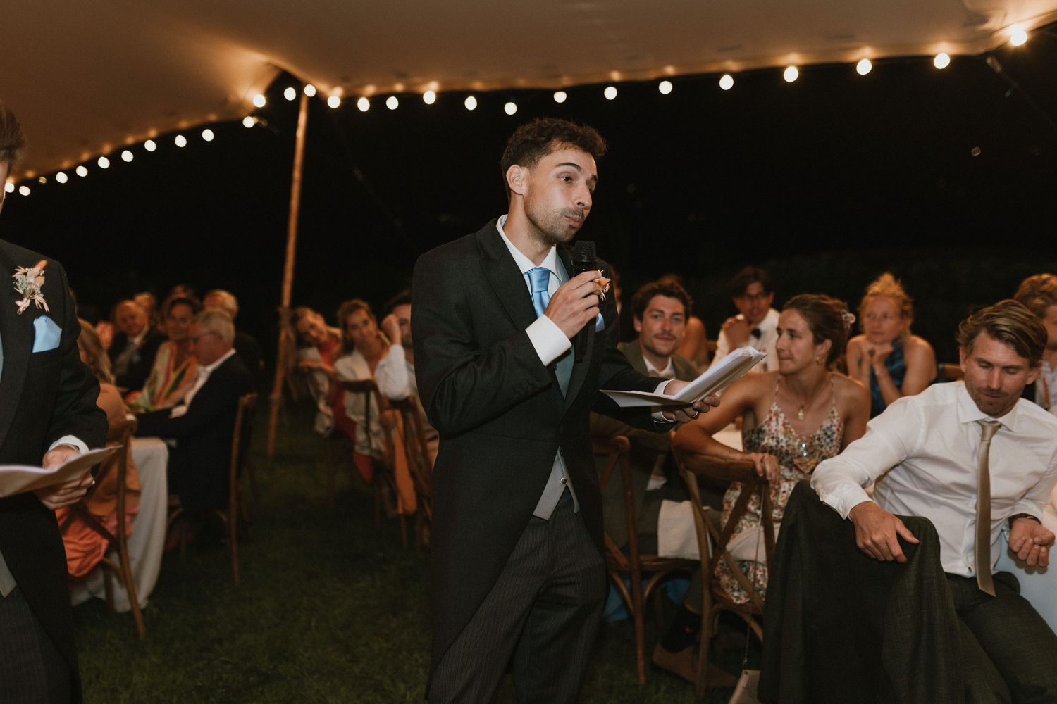 Wedding reception under tent with speaker entertaining guests during evening celebration.