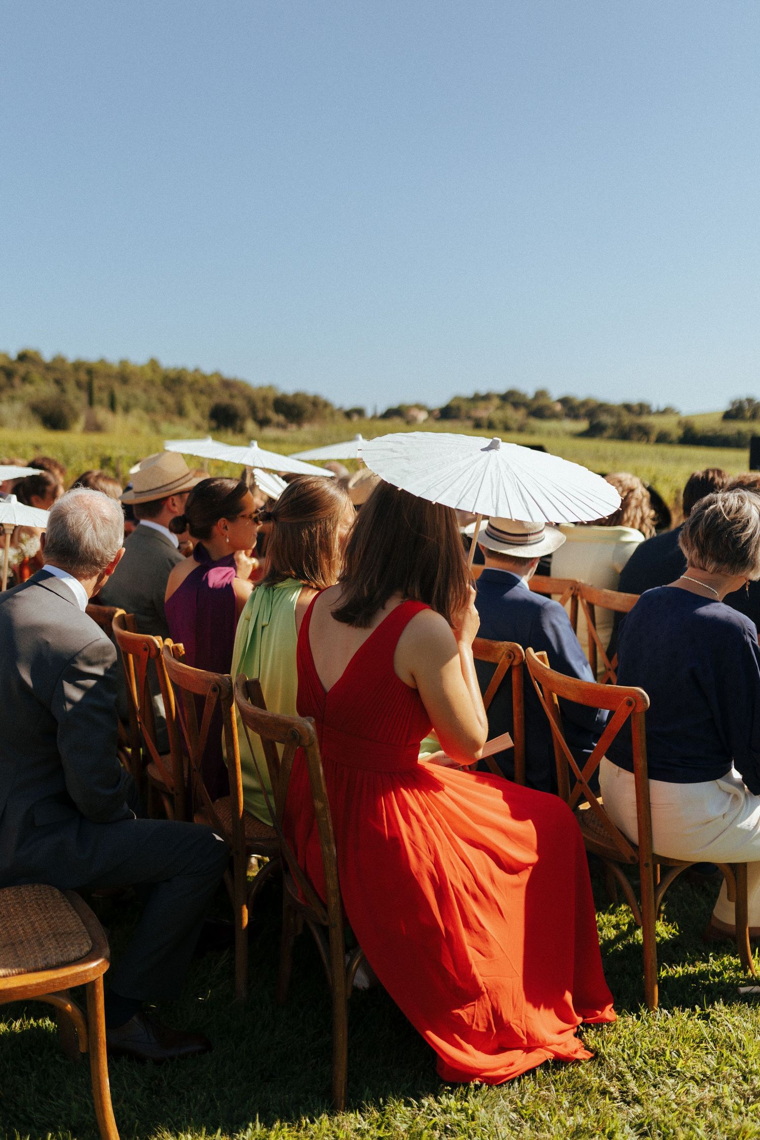 Guests at an outdoor event holding white parasols while sitting in wooden chairs on a grassy lawn under blue skies.
