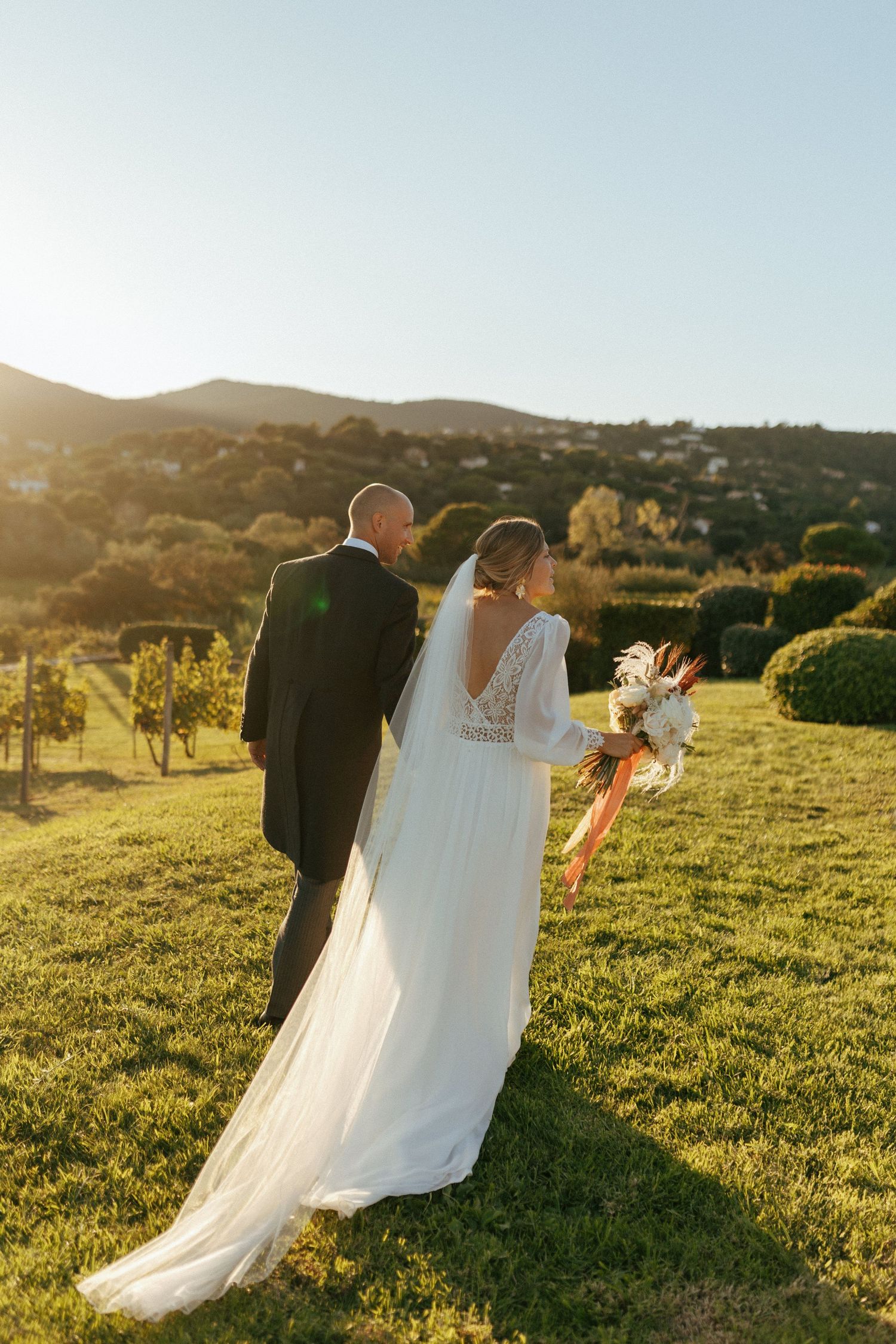 A couple in wedding attire stands in a scenic vineyard during golden hour with flowing white dress.