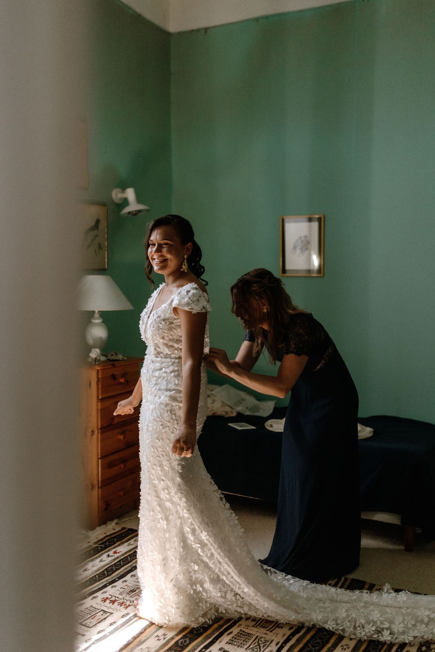 A bride in a lace wedding dress stands while someone in navy helps with final adjustments in a mint green room.