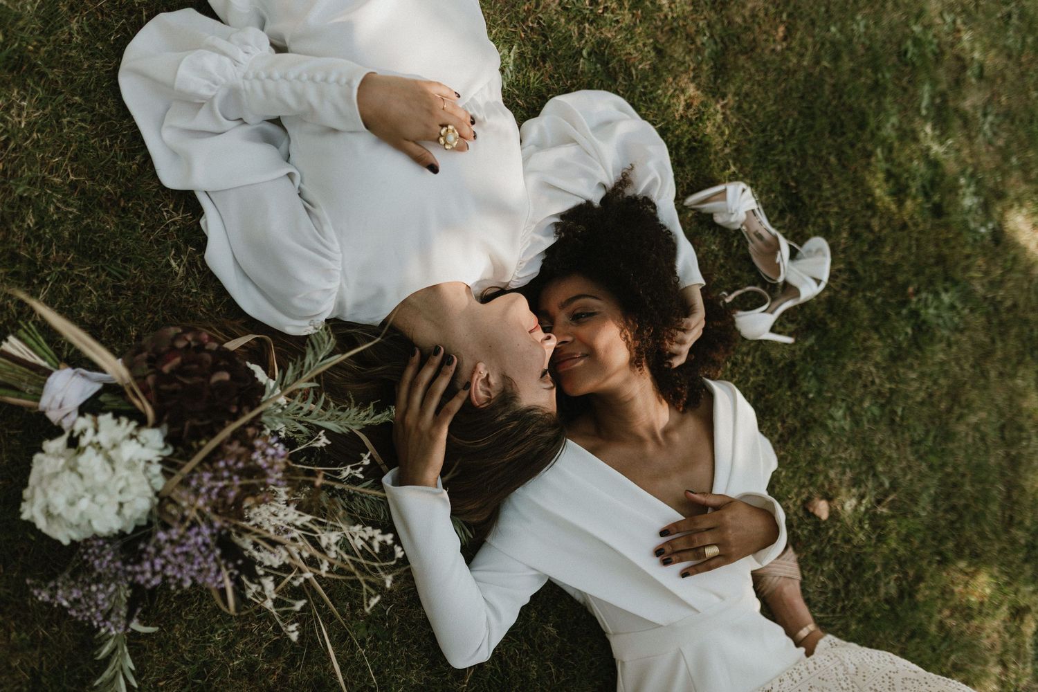 A couple in white outfits laying on grass with flowers during a wedding photoshoot.