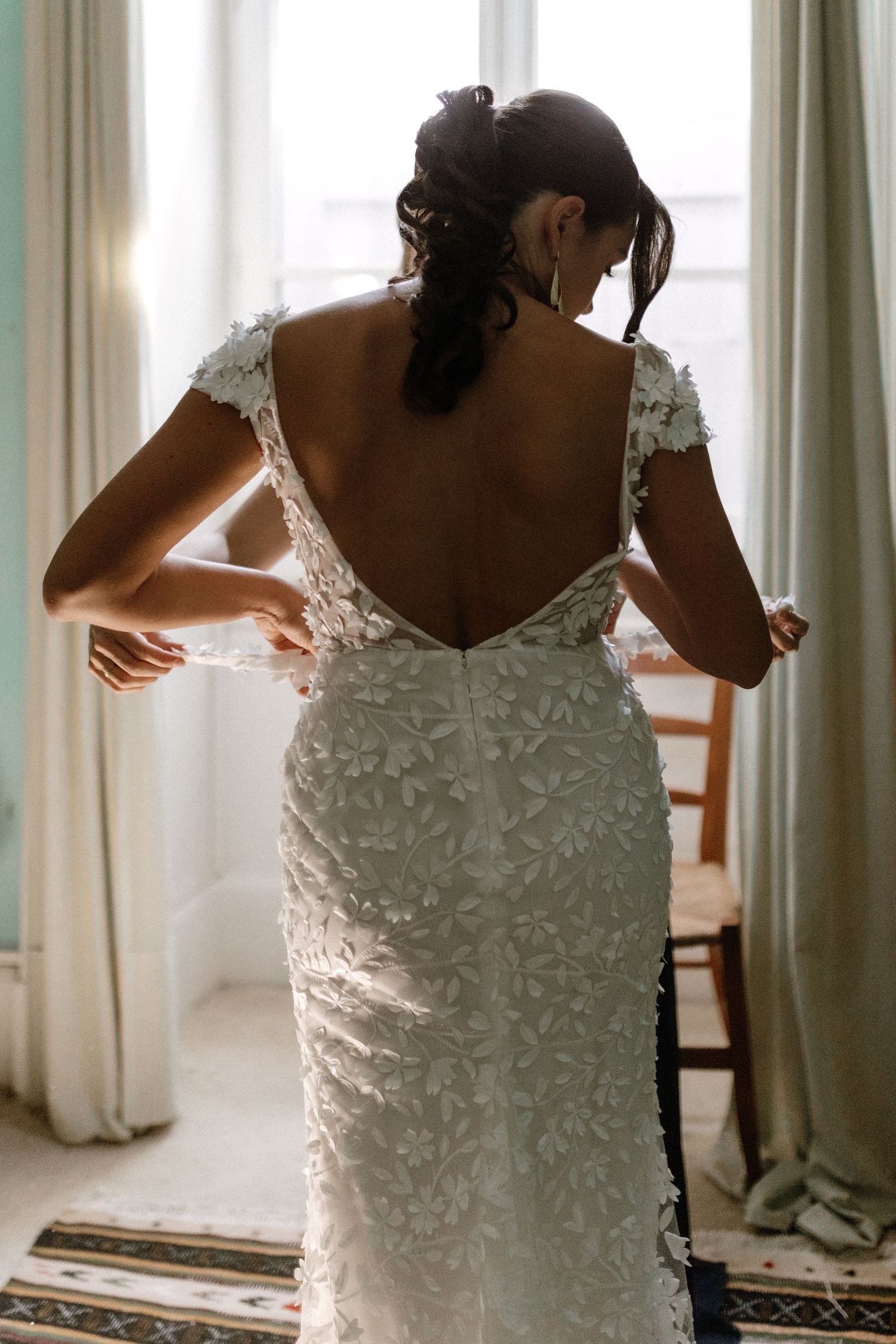 Back view of a bride wearing an intricate lace wedding dress with cap sleeves against window light.