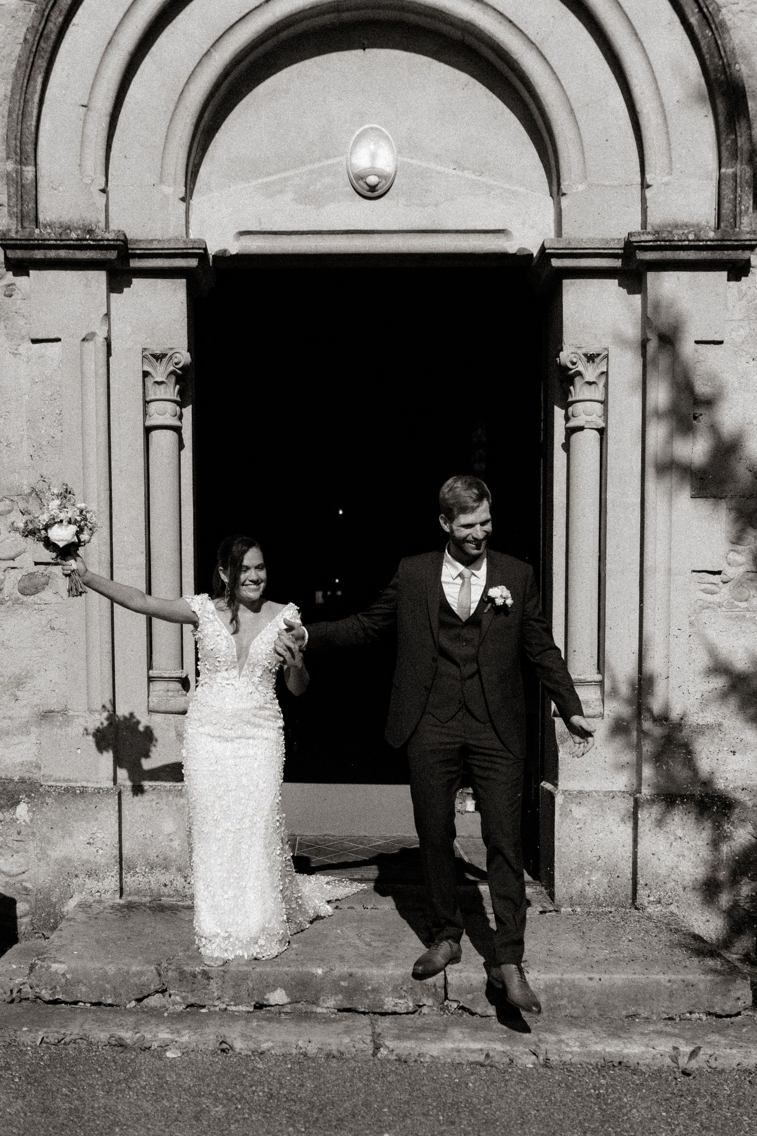 A joyous couple exits a historic church doorway in black and white, celebrating their wedding day.