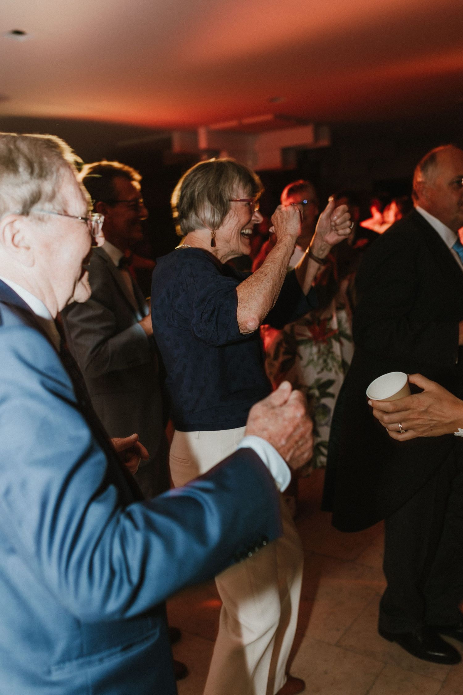 Wedding guests dance and celebrate at an indoor reception with dramatic red lighting.