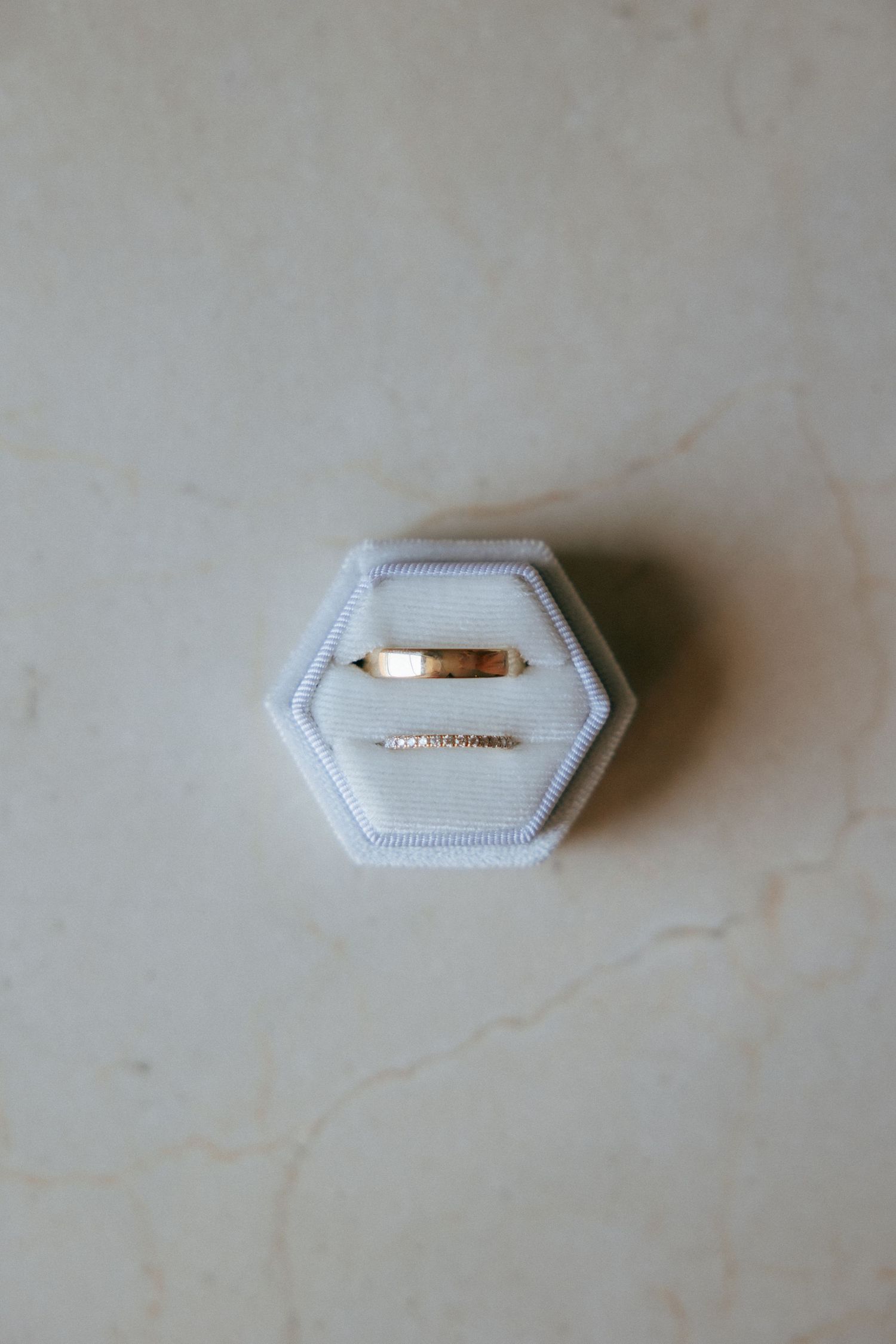 A delicate gold wedding ring sits in a white hexagonal ring box on a light marble surface.