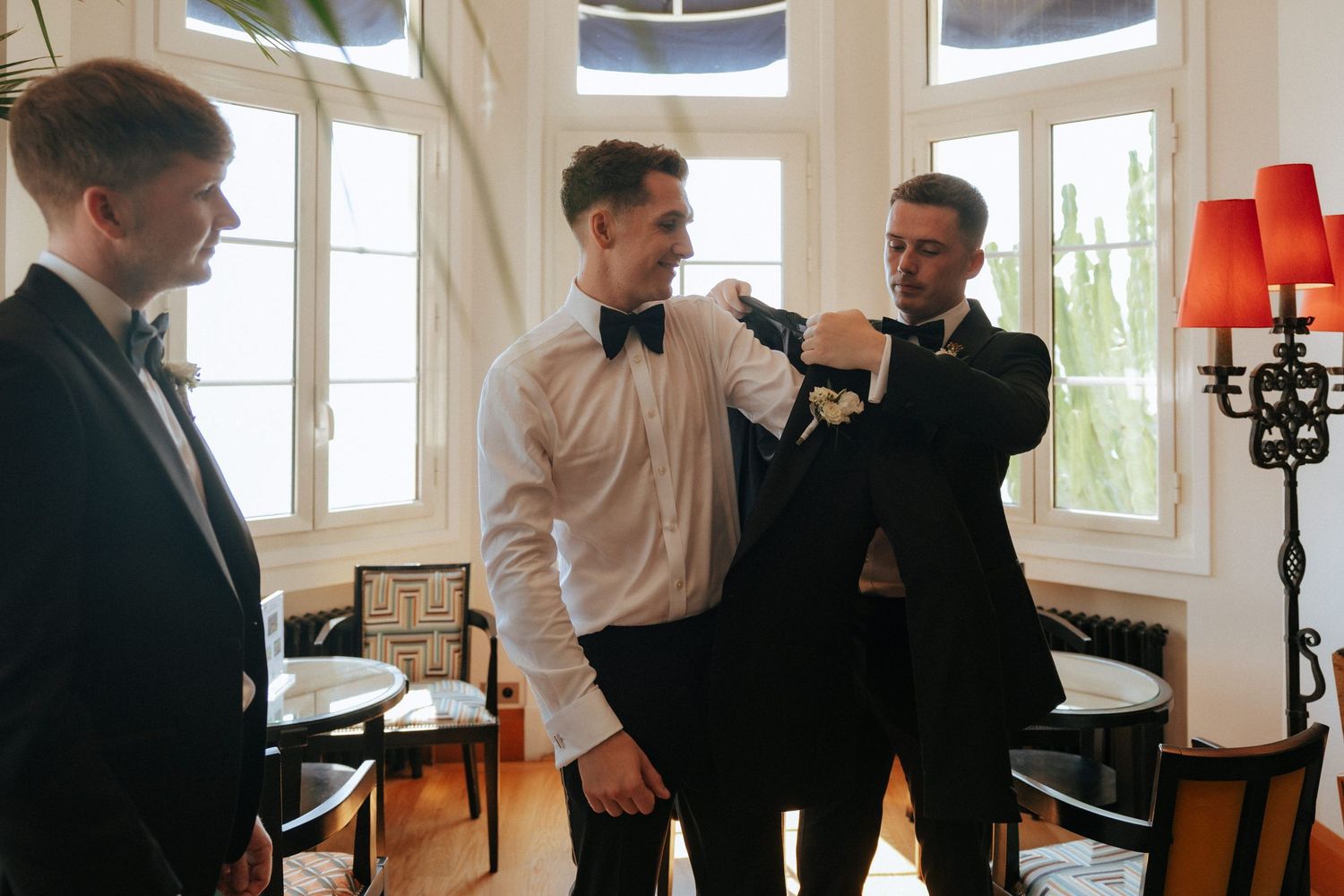 A wedding day moment showing final preparations with formal attire adjustments in a bright room with tall windows.