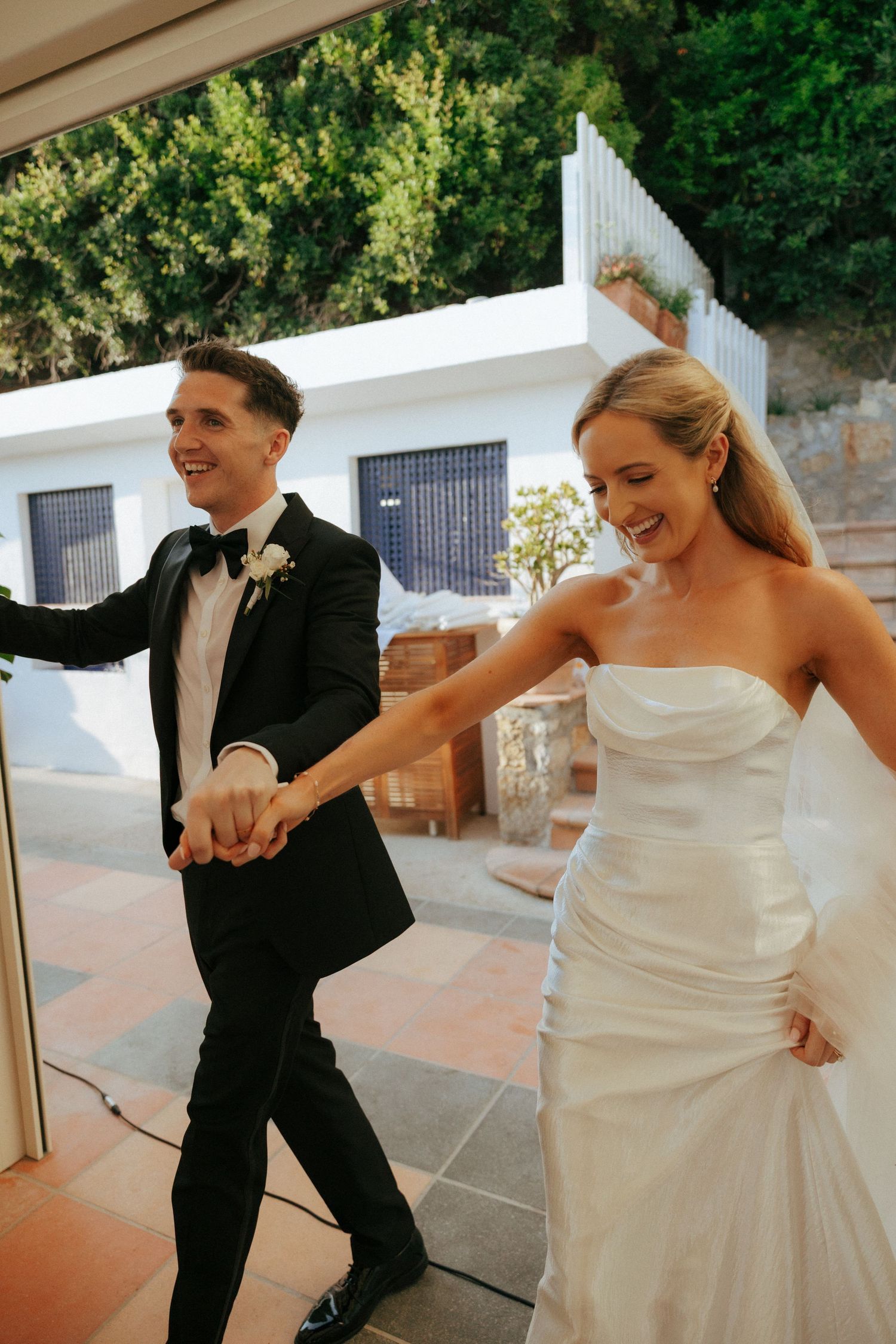 A wedding couple shares a joyful moment dancing together on a stone patio with white walls in the background.