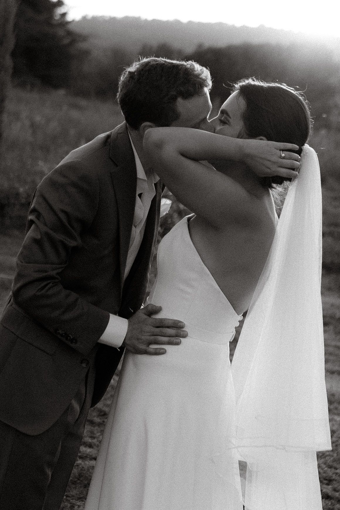 A romantic black and white wedding photograph of a couple sharing their first kiss in an outdoor setting with soft sunlight.