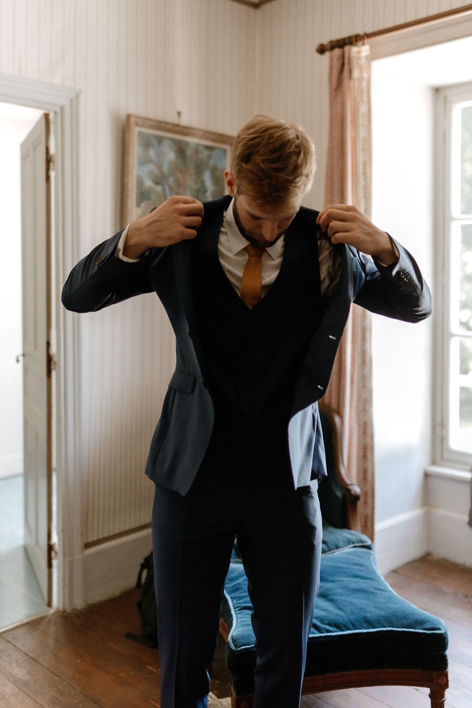 A person adjusts their formal attire while getting ready in a sunlit room with hardwood floors.