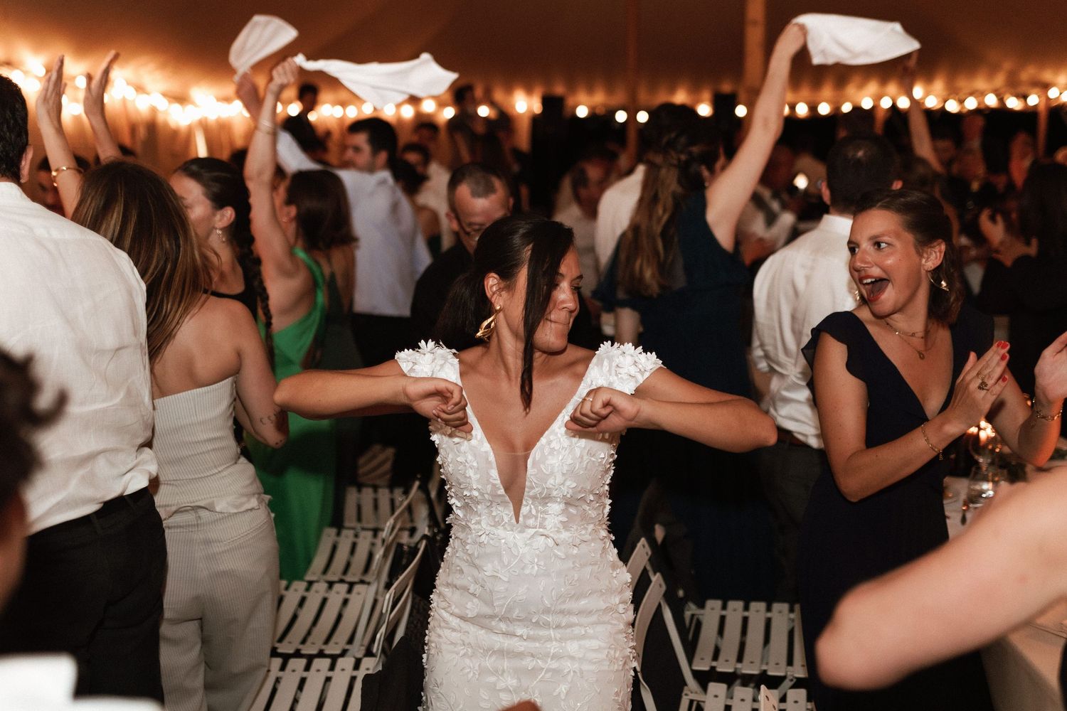 Wedding reception dance party with a crowd of guests waving napkins and dancing under string lights.