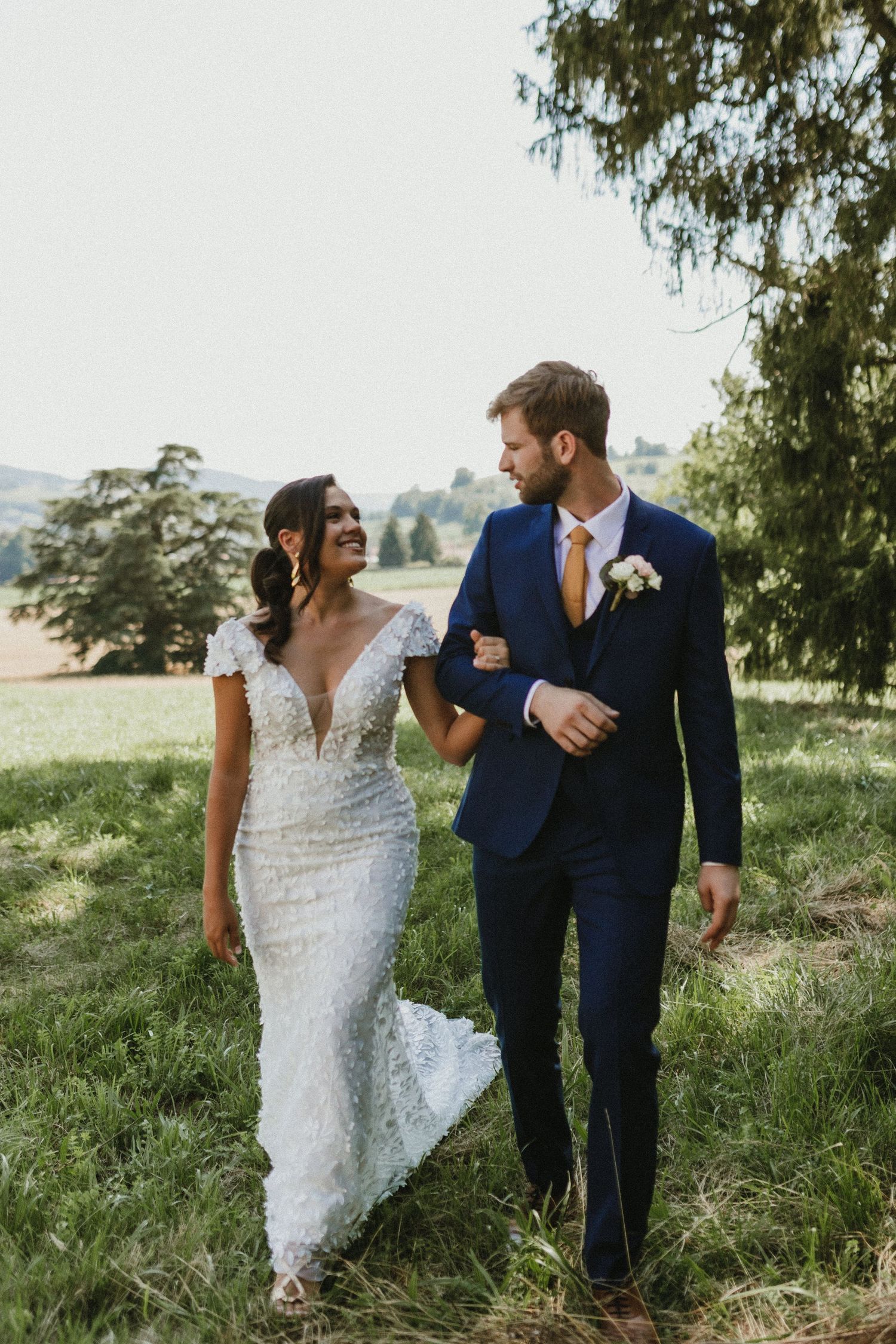 Newlywed couple in elegant wedding attire smiling at each other while walking through a grassy hillside.