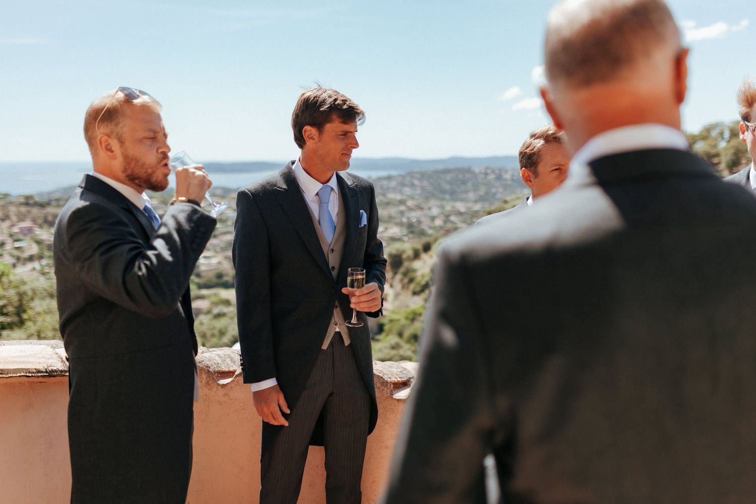 Men in formal attire conversing on outdoor terrace with scenic mountain view in background.