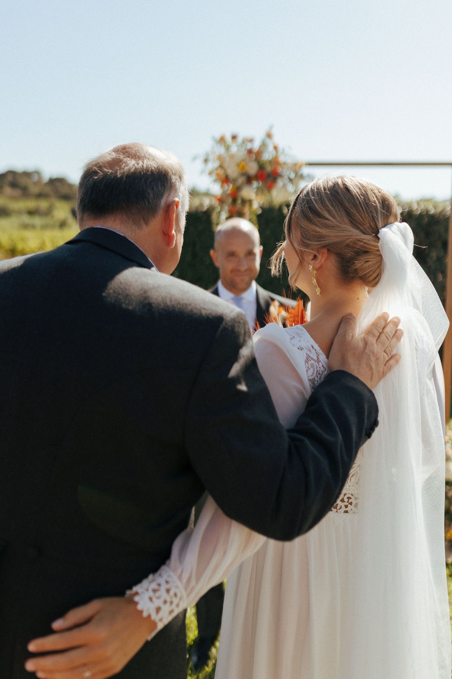 An outdoor wedding ceremony with a bride in white lace and her escort walking towards the officiant.