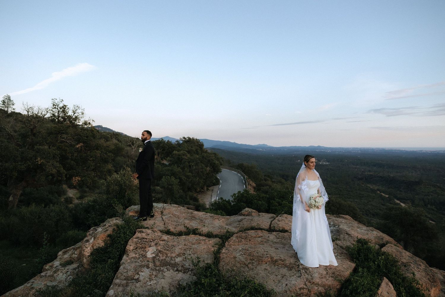 Two people standing on rocky mountaintop overlook with scenic valley vista at sunset.