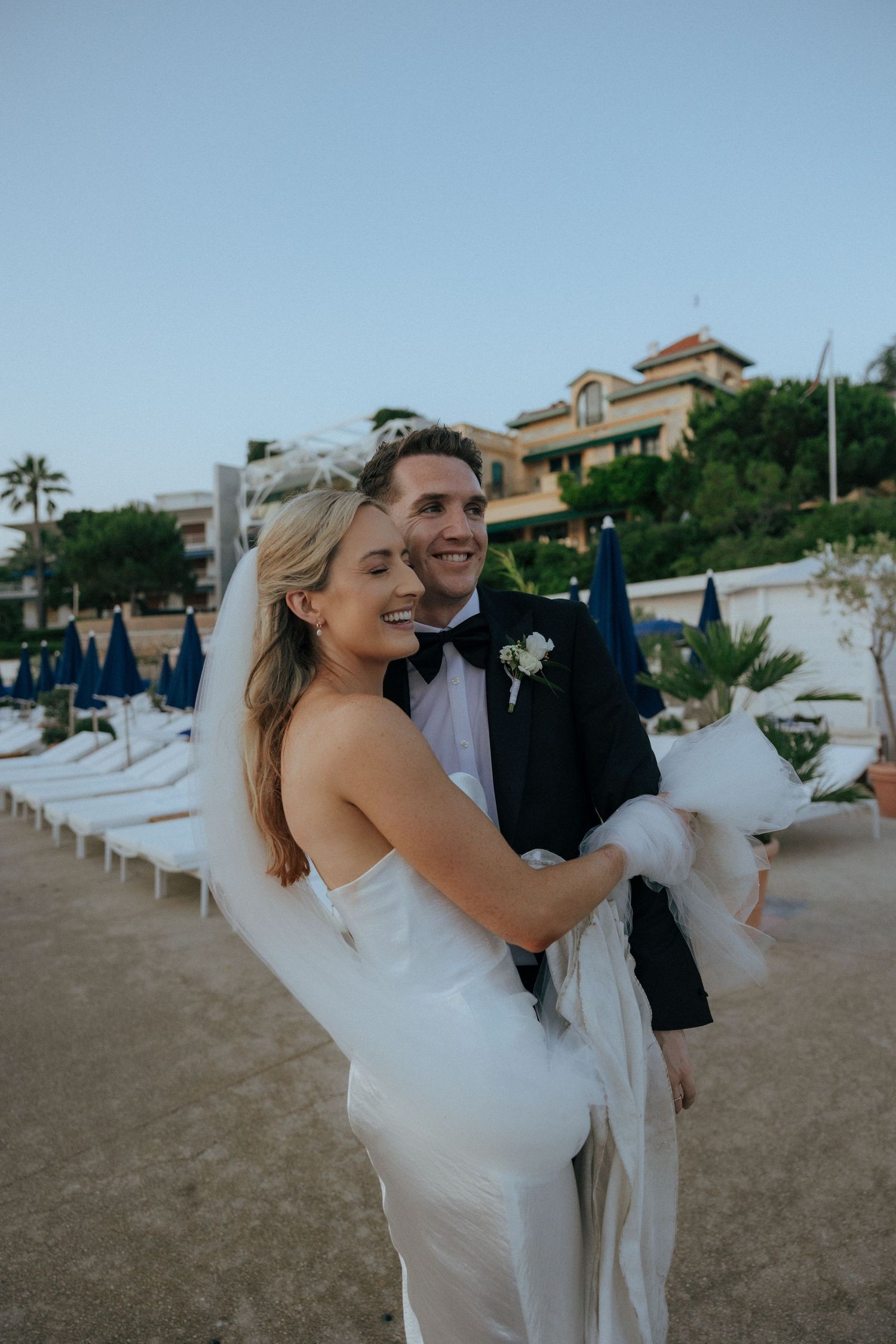 A couple in wedding attire poses on a beach with lounge chairs and palm trees in the background.
