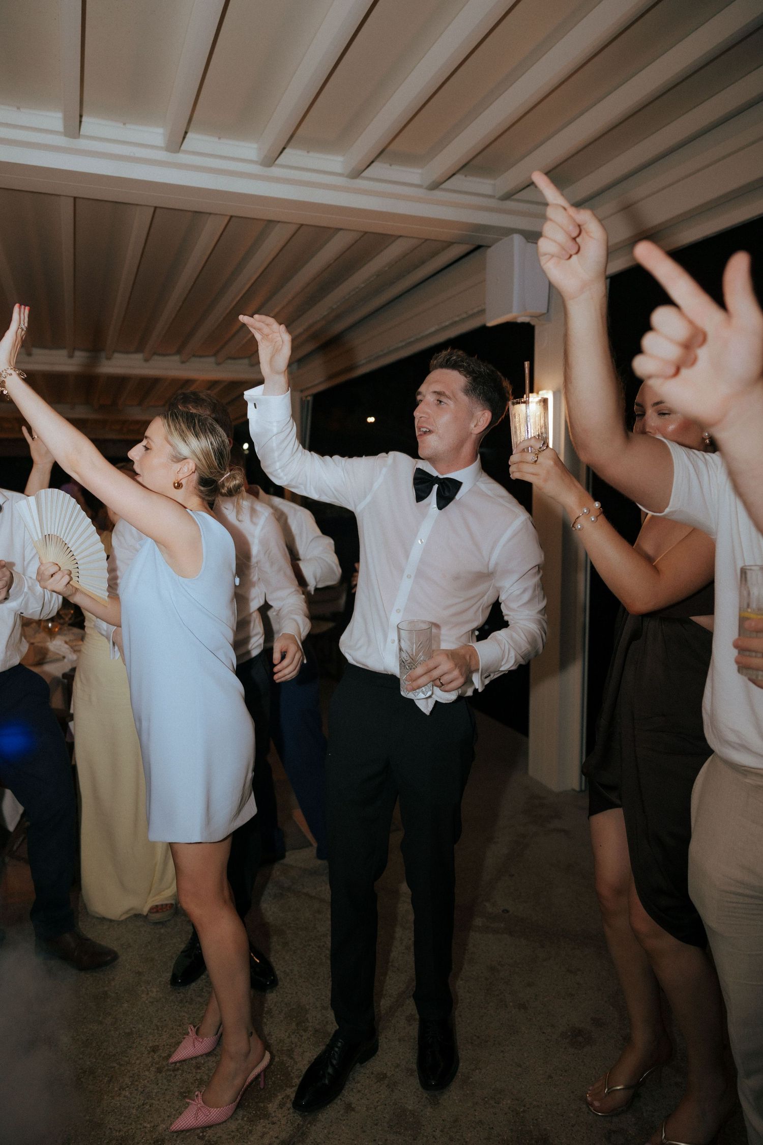 Party guests dancing with arms raised under white ceiling beams at a formal celebration.