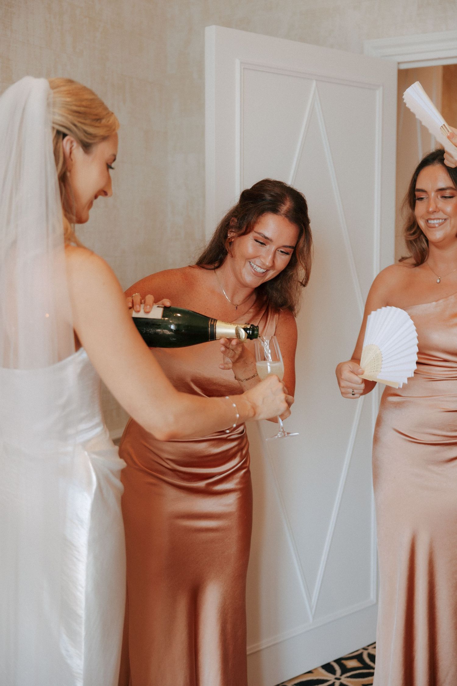 Three smiling people in formal attire share a celebratory moment, with champagne and fans at a special event.