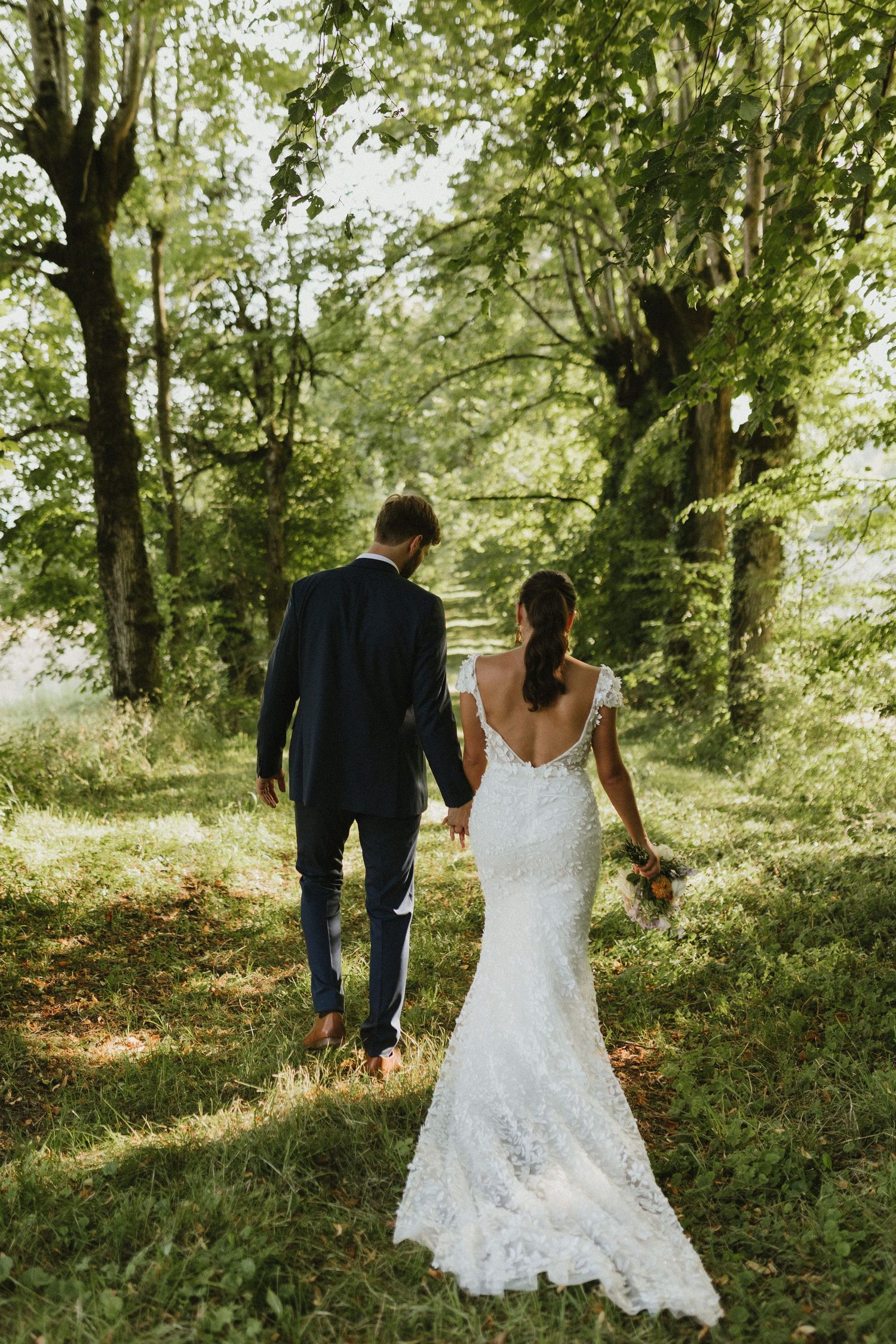 A couple walks through a sunlit forest path, the bride wearing a stunning backless lace wedding gown.