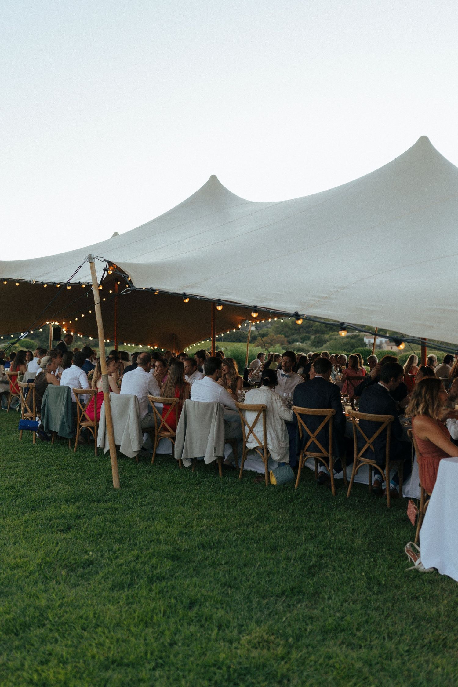 Long dining tables under an elegant white tent canopy at an outdoor wedding reception with string lights.