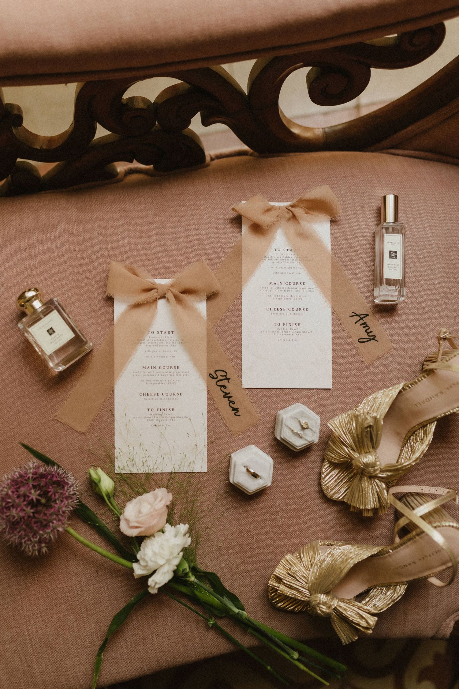 Wedding invitation cards with beige bows displayed alongside perfume bottles, flowers, and golden metallic decorative elements.