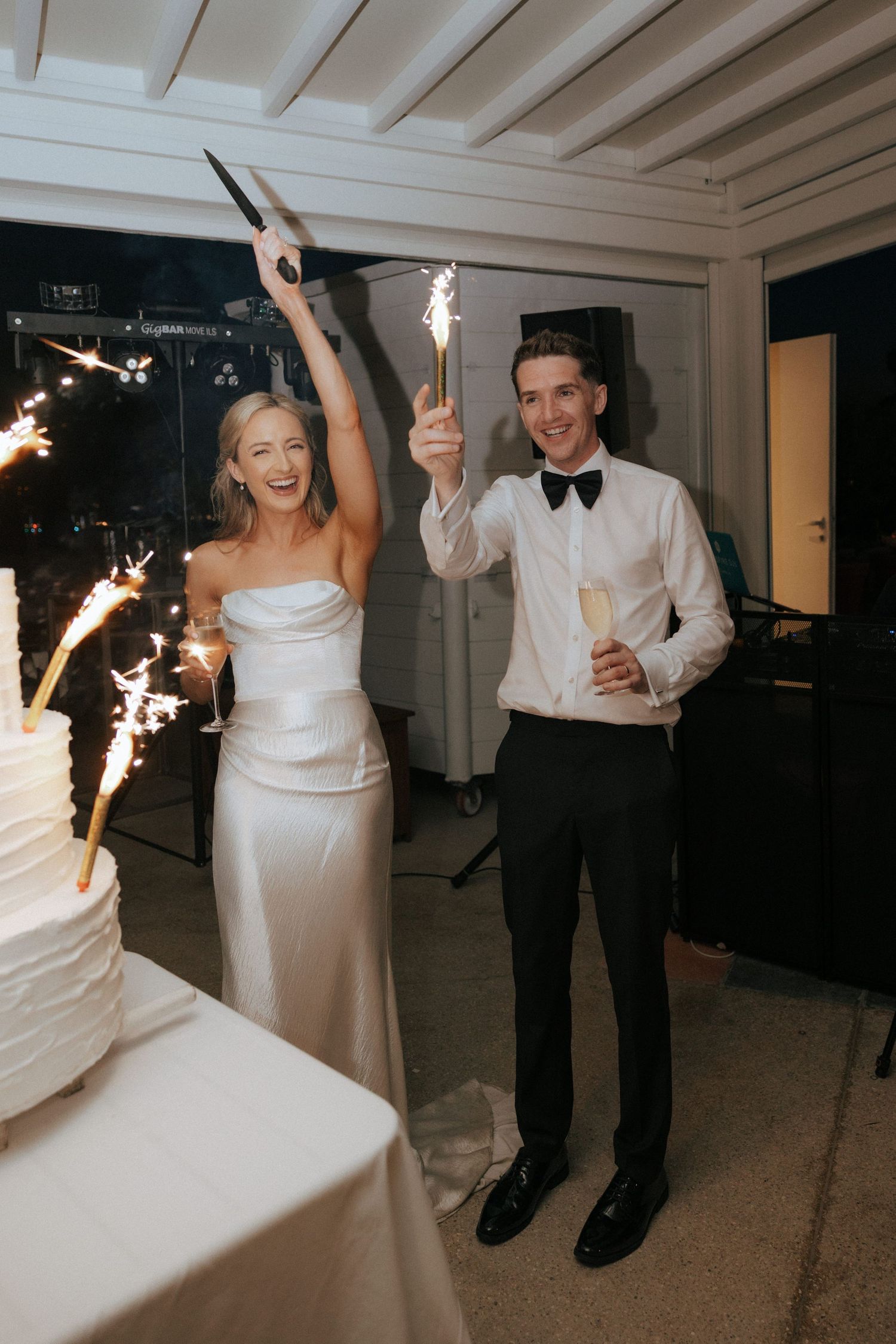 Newlyweds celebrate at their wedding reception with sparklers beside their illuminated white wedding cake.