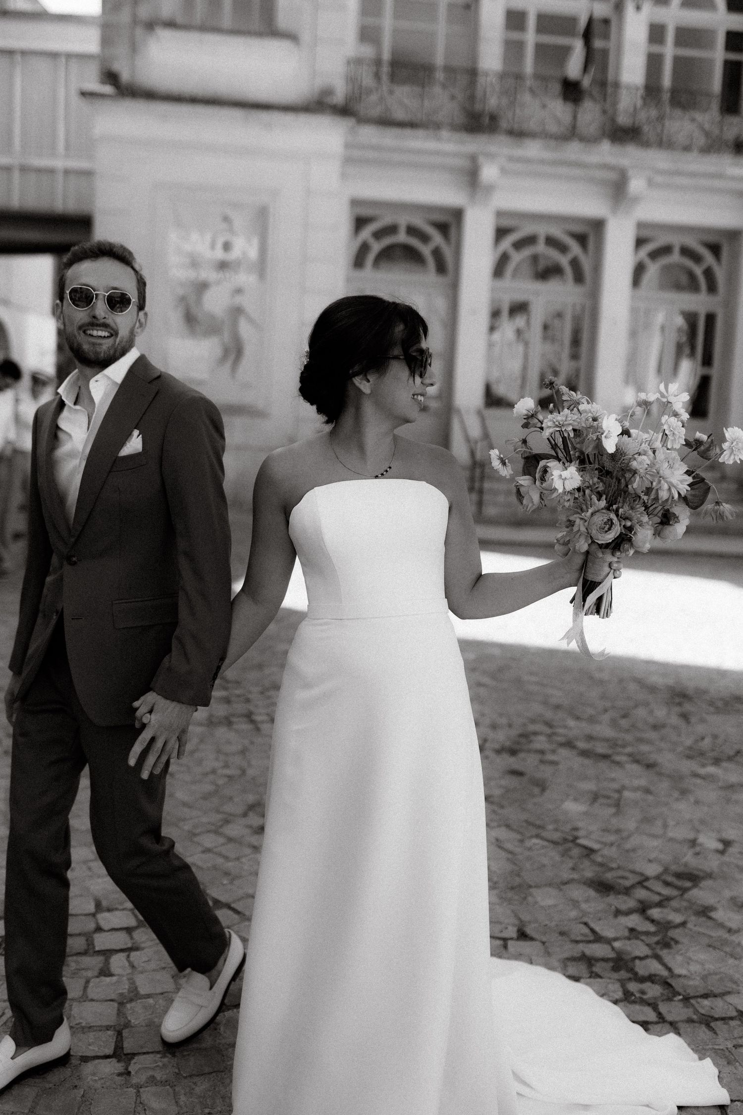 Black and white wedding photo showing couple in formal attire with bride holding bouquet on cobblestone street.