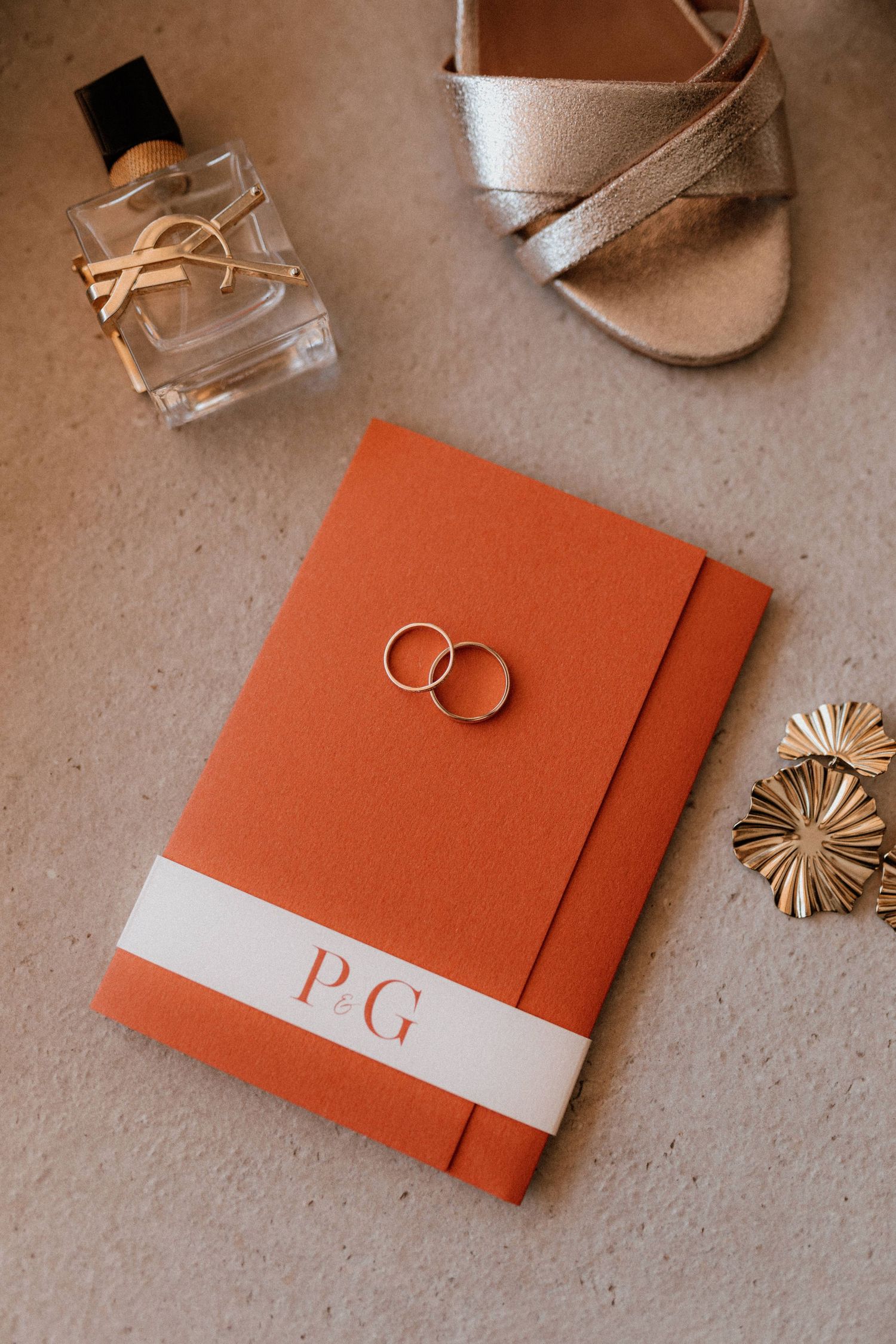 Wedding details featuring a perfume bottle, silver sandal, orange monogrammed book and gold wedding rings on neutral surface.