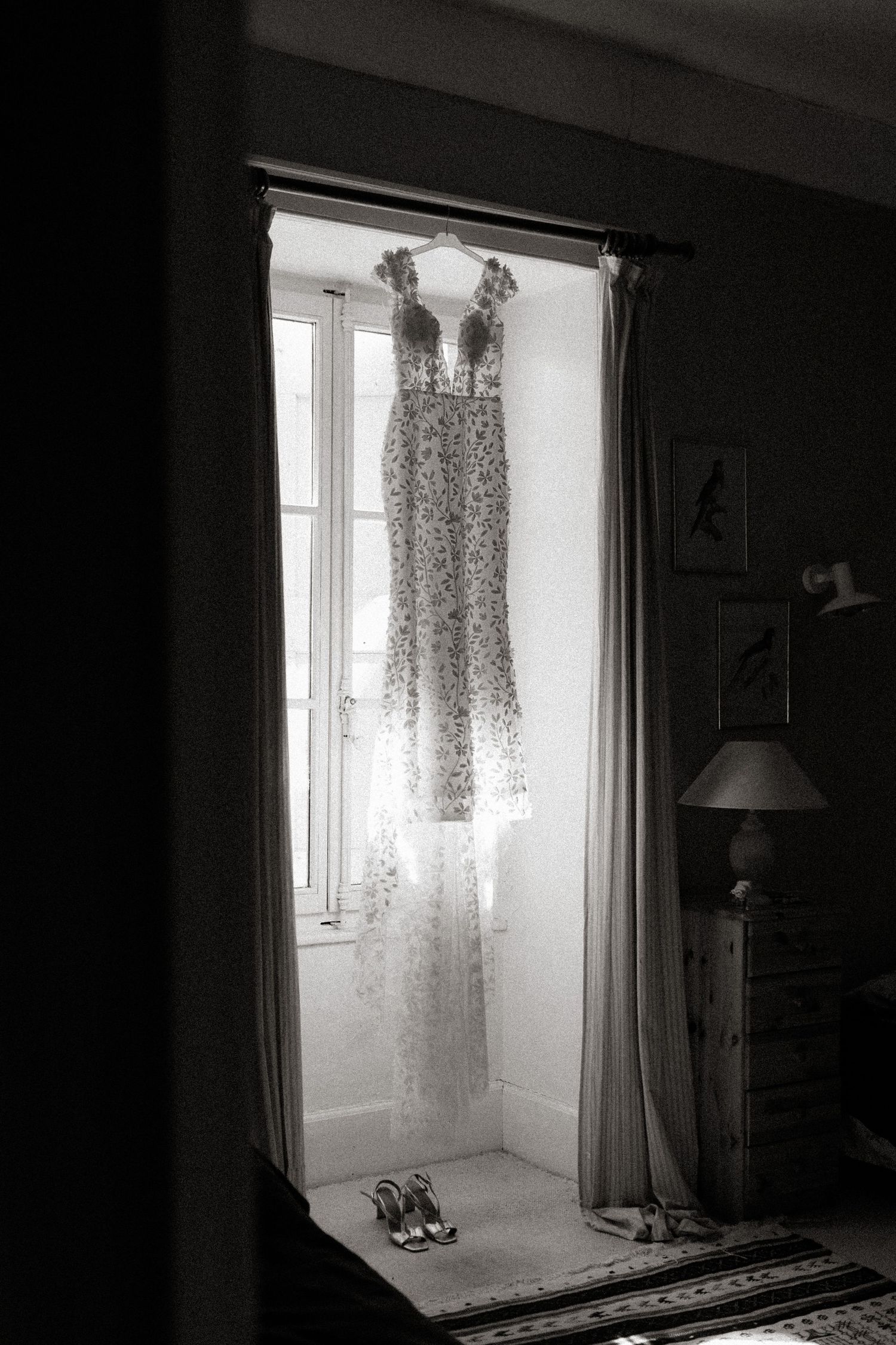 A delicate lace dress hangs in silhouette by a sunlit window with a pair of shoes placed beneath it.