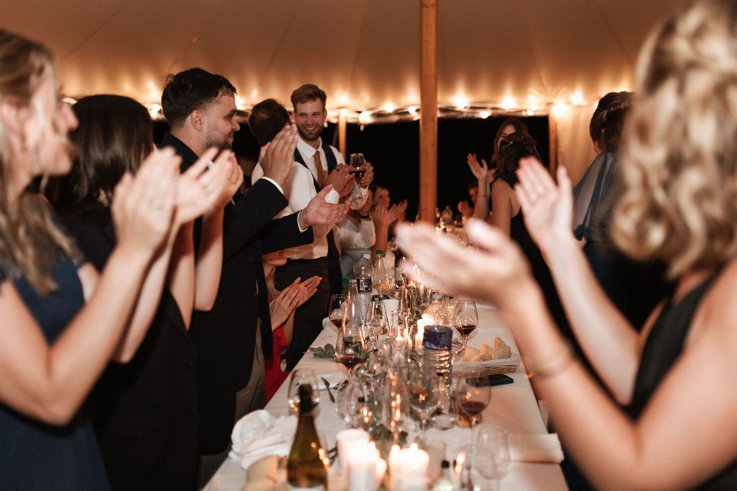 Wedding guests clapping and celebrating at long tables under string lights in a tent.