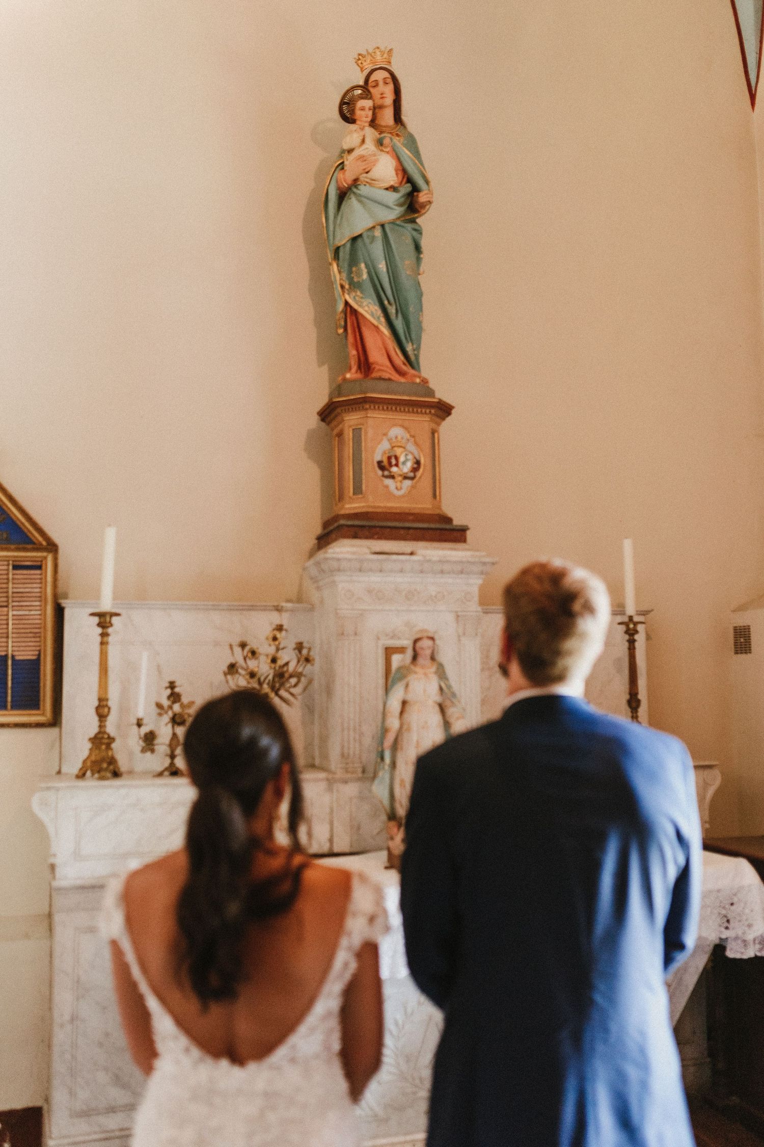 A couple stands before a religious statue and altar during a wedding ceremony in a church.