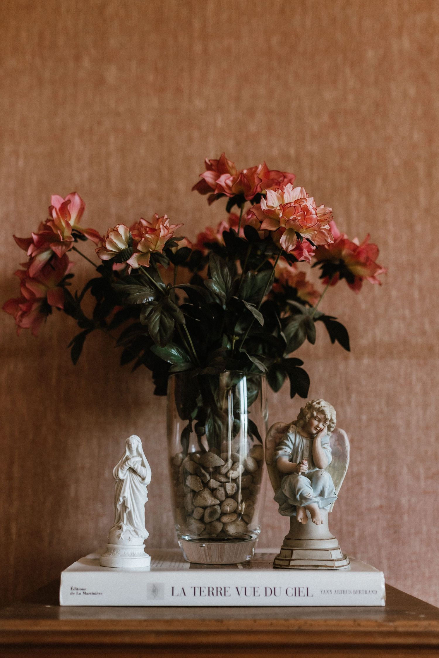 Pink flowers in a clear vase with decorative stone filling sit atop a book with classical figurine statues.