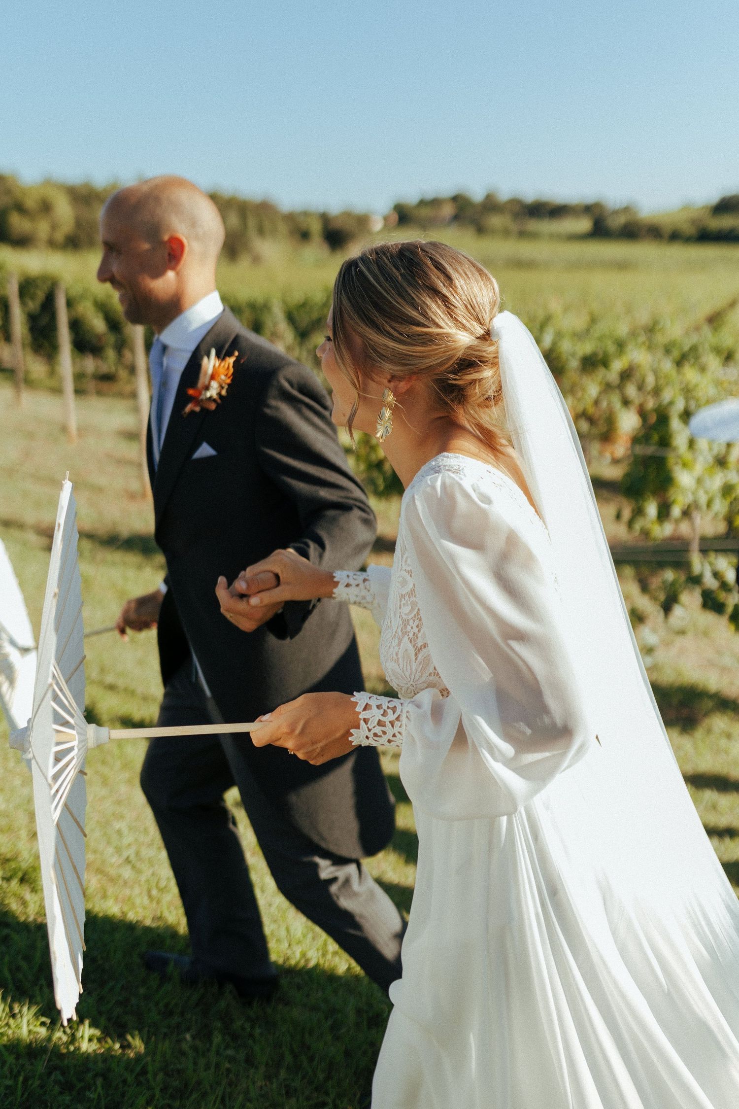 A couple with parasols celebrate their wedding ceremony in a sunlit vineyard setting.