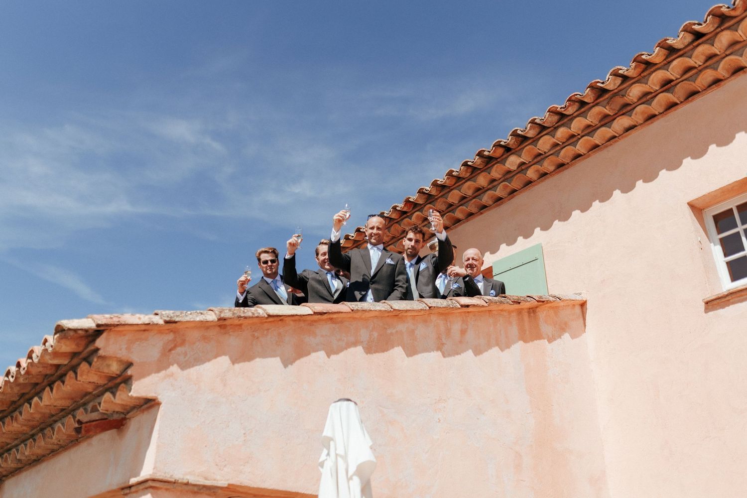 People in formal attire gather on a terracotta-roofed Mediterranean building against a blue sky background.