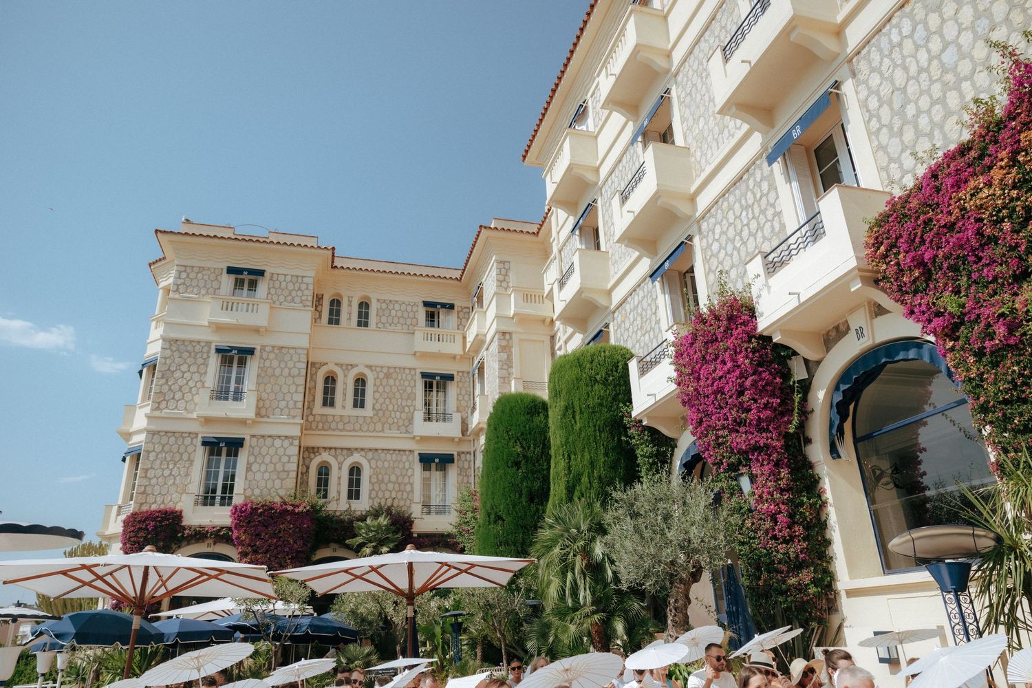 Mediterranean luxury hotel with elegant balconies, pink bougainvillea flowers, and manicured topiary shrubs against beige walls.