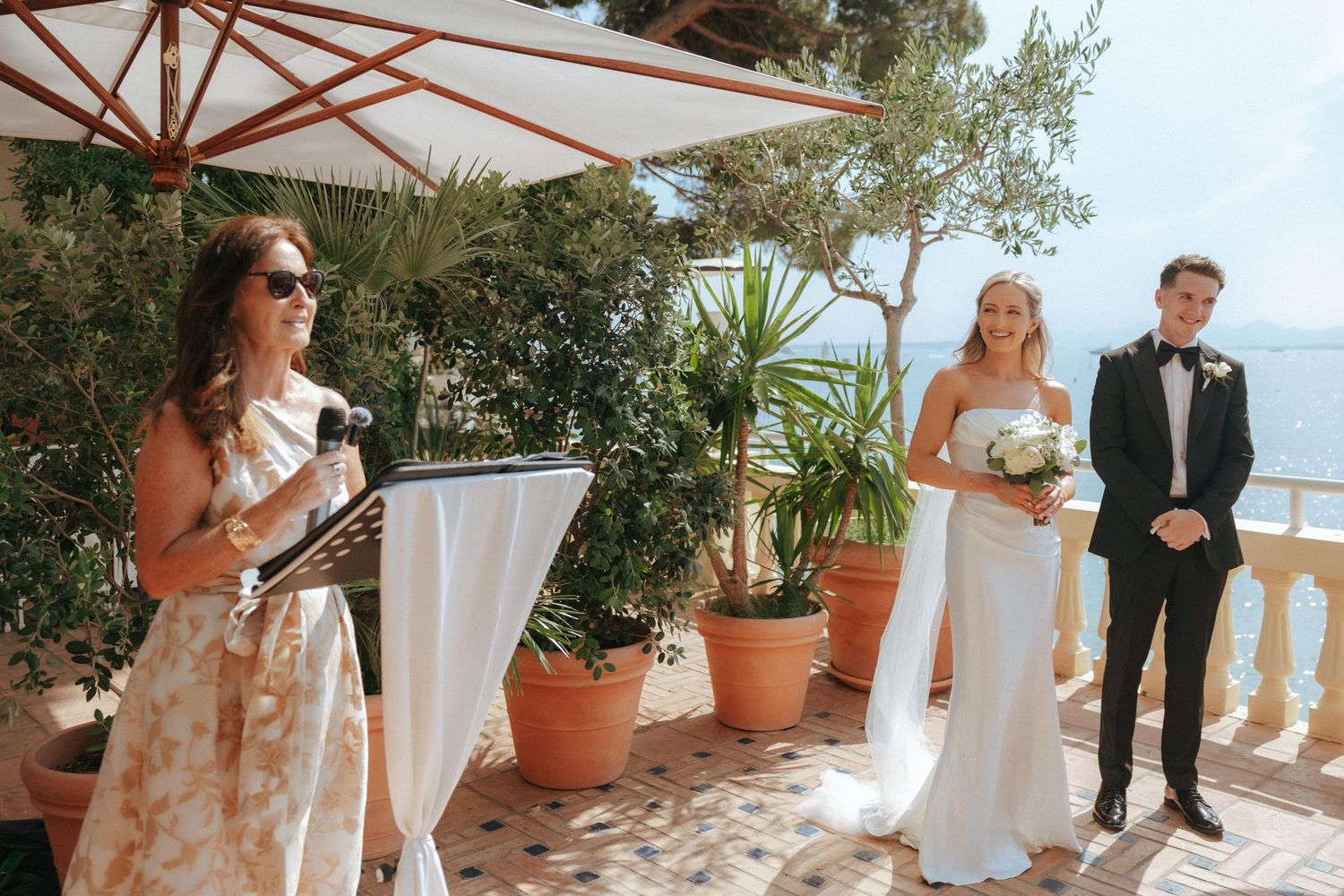 An outdoor ceremony takes place on a Mediterranean terrace with potted plants and a white umbrella overlooking the ocean.