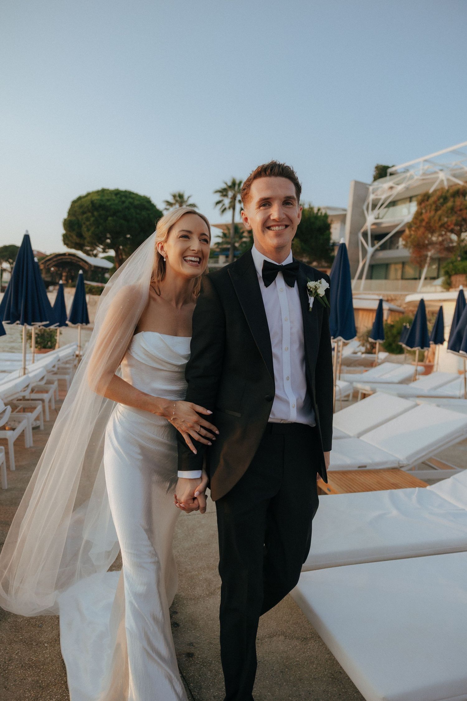 A joyful wedding couple pose together on a sunny beach terrace with palm trees and lounge chairs in the background.