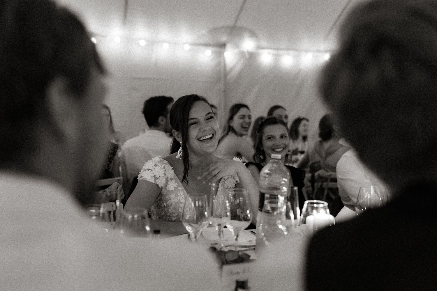 Wedding reception guests share joyful laughter under string lights inside an elegant tent setting.