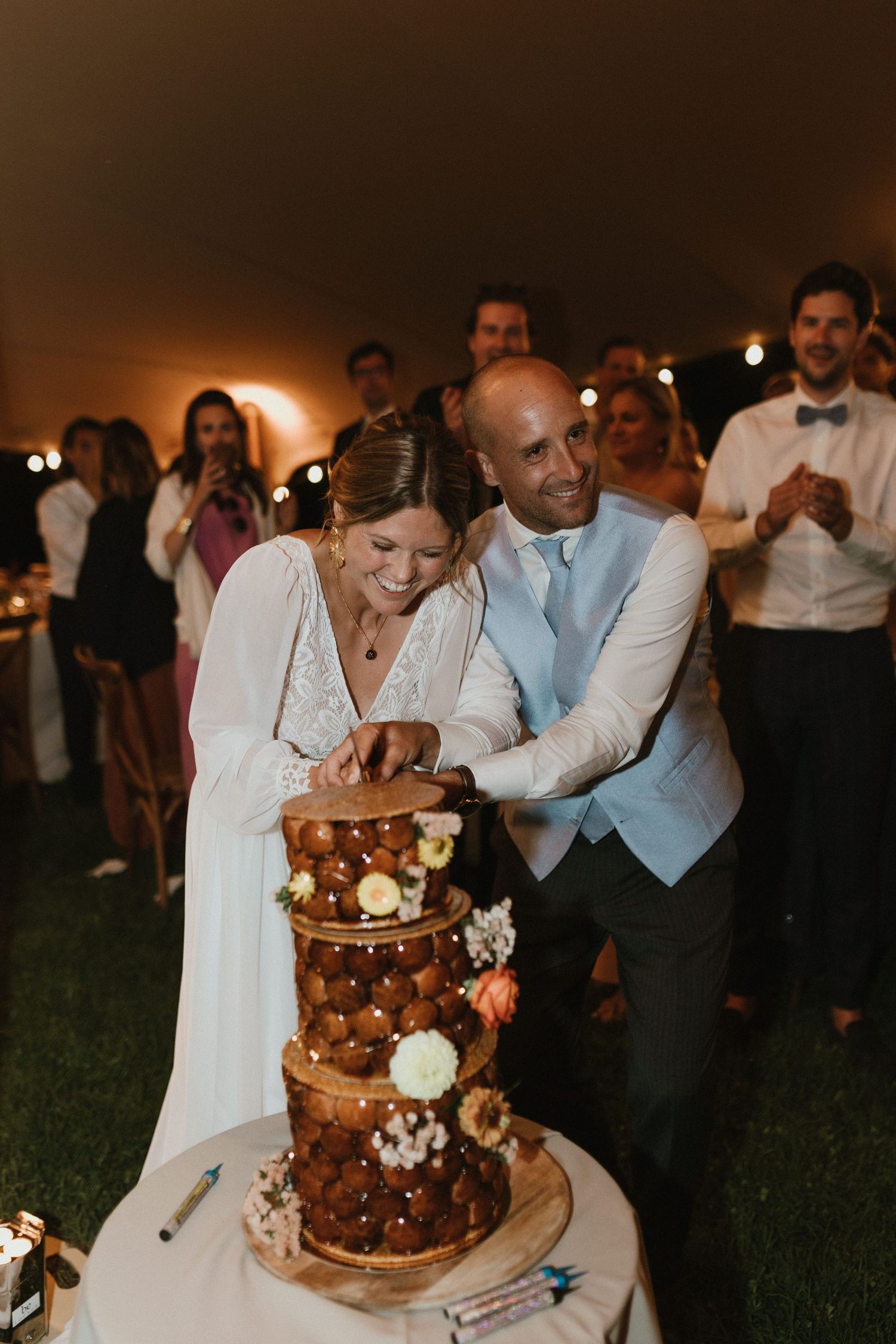 A couple cutting their elegant chocolate wedding cake adorned with flowers and fairy lights at an outdoor evening reception.
