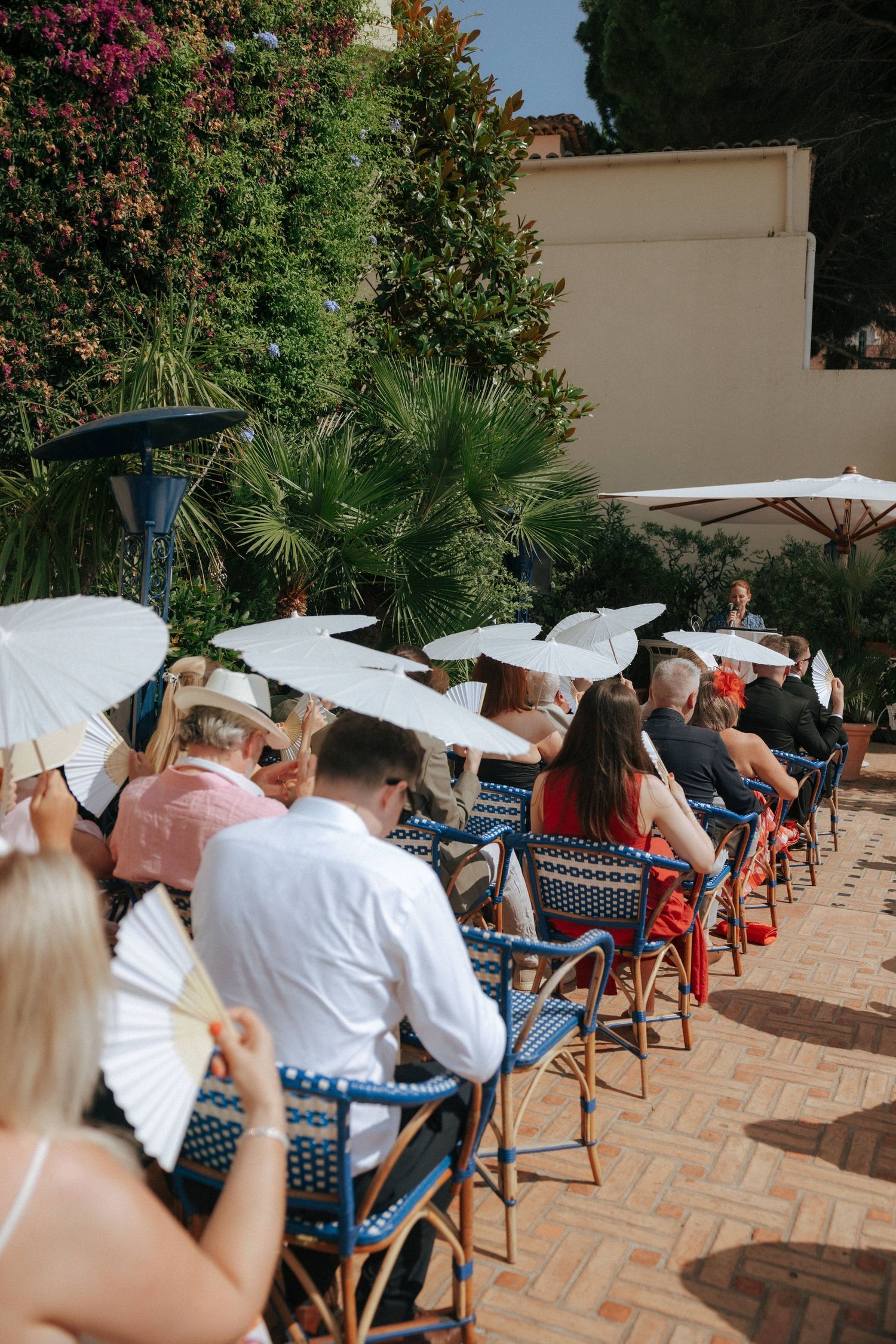Wedding guests seated in blue chairs holding white parasols at an outdoor venue.