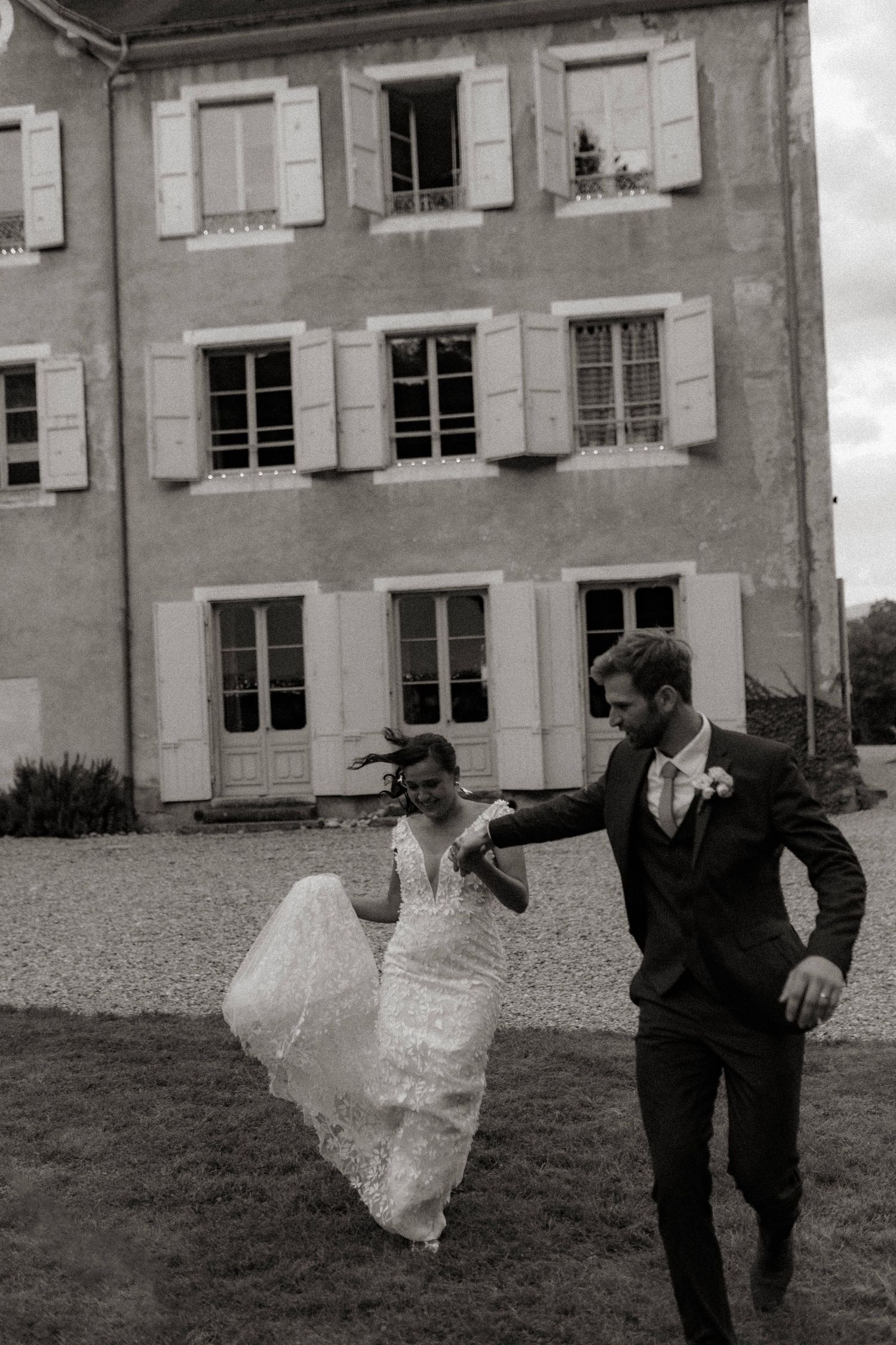 A joyful couple in wedding attire dances together on the lawn in front of a French chateau with white shuttered windows.