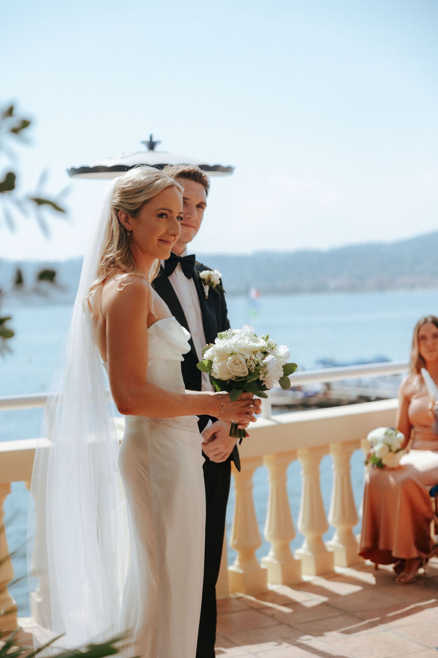 Outdoor seaside wedding ceremony with couple exchanging vows on a white balcony overlooking the ocean.