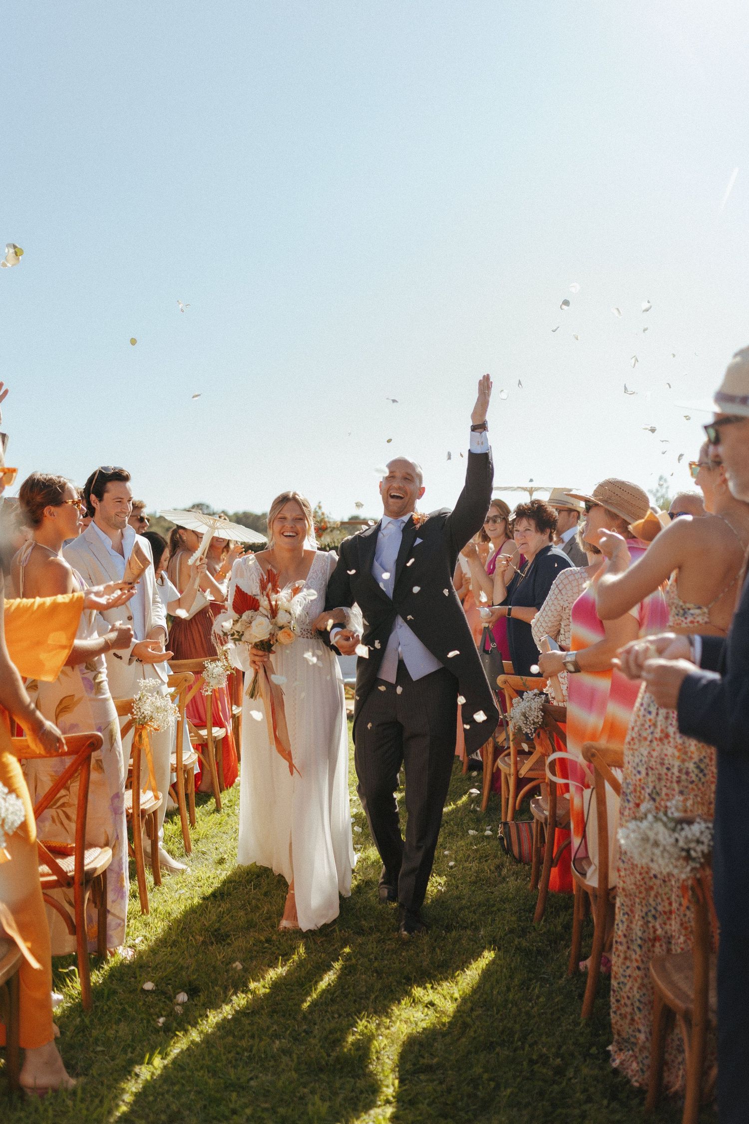 Joyous wedding couple walks down aisle while guests throw confetti in golden afternoon sunlight.