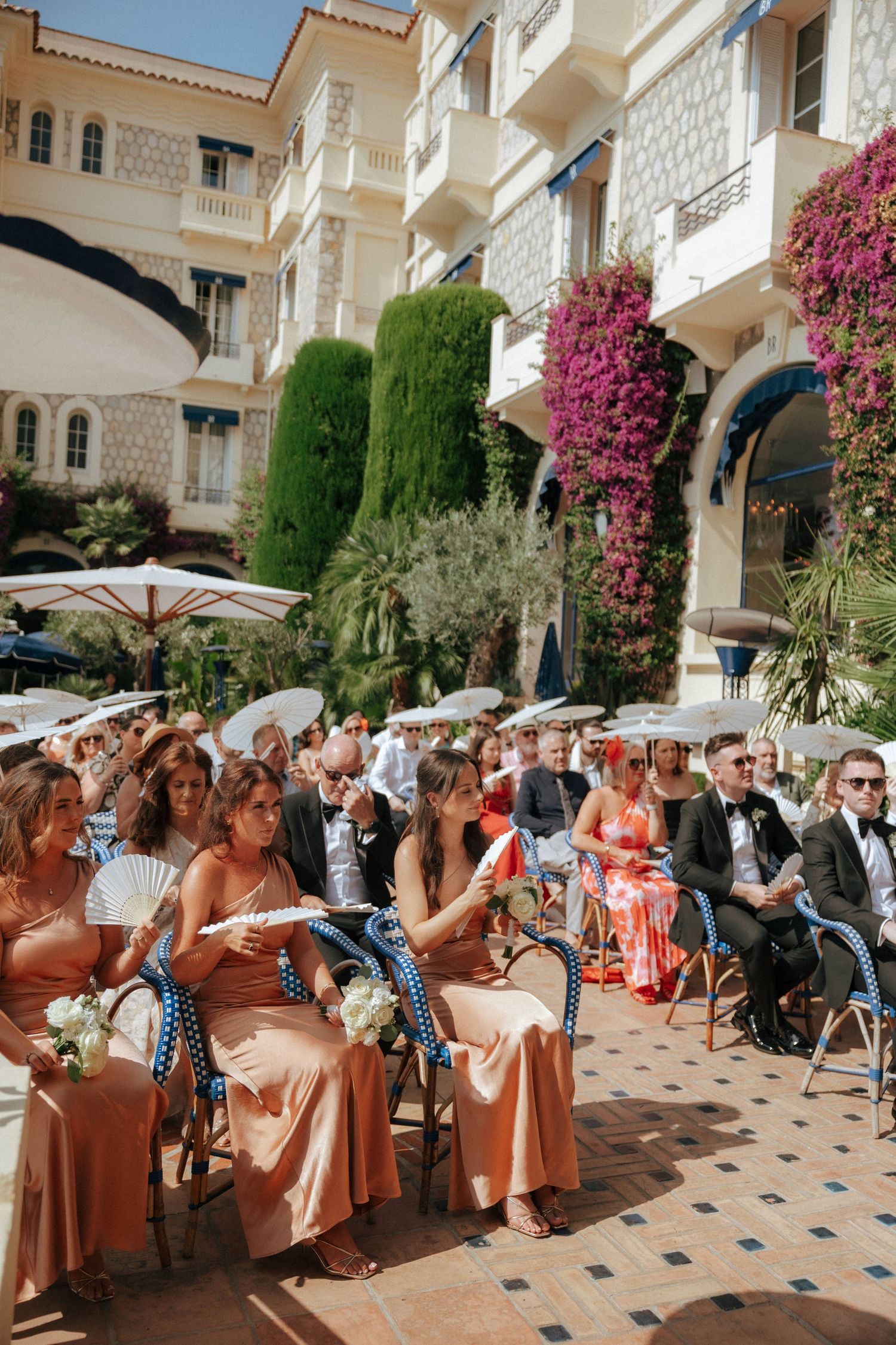 Wedding guests seated at outdoor tables with pink floral decor and Mediterranean architecture.